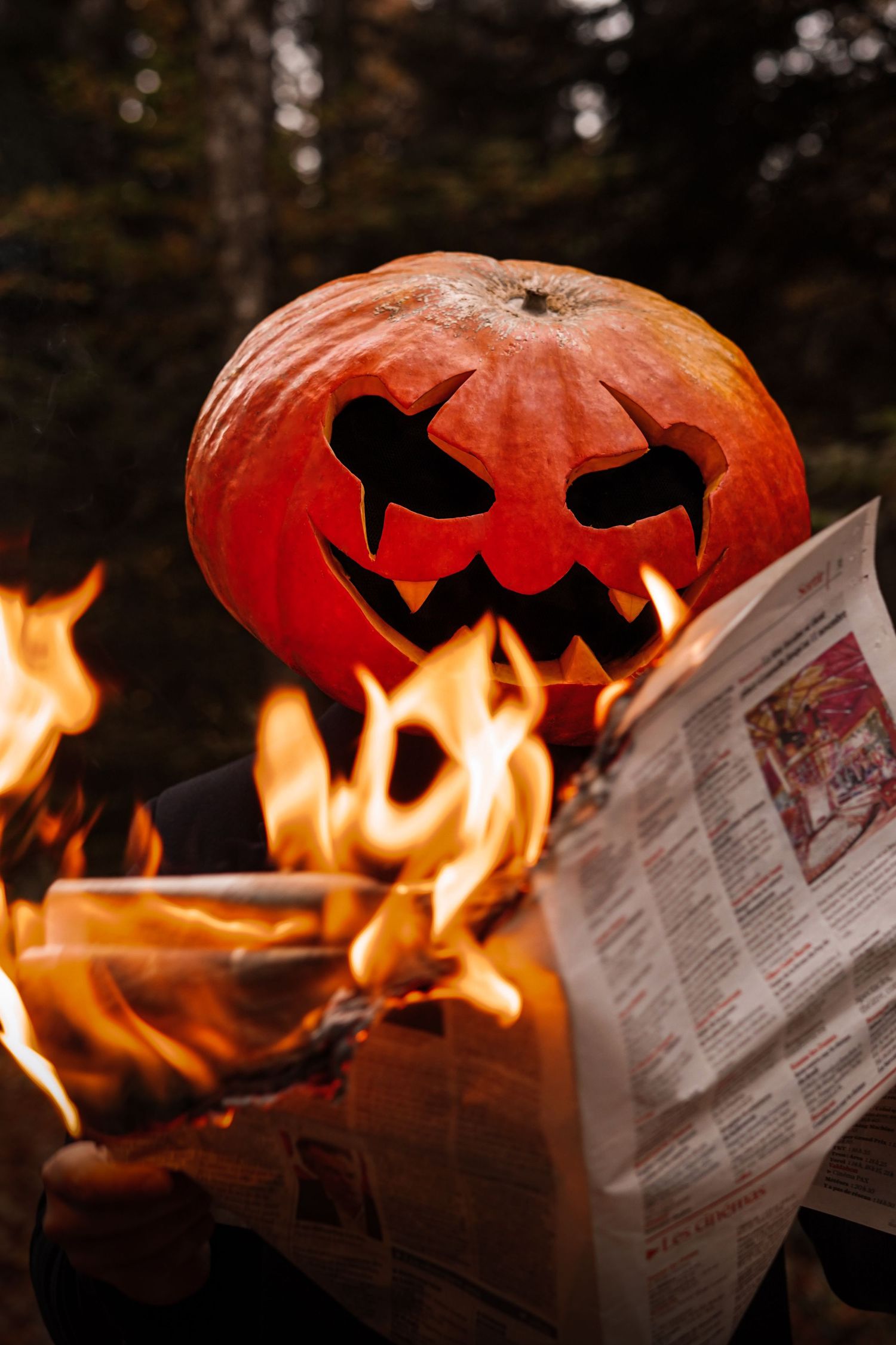 Carved Halloween jack-o-lantern reading a burning newspaper in a dark forest.