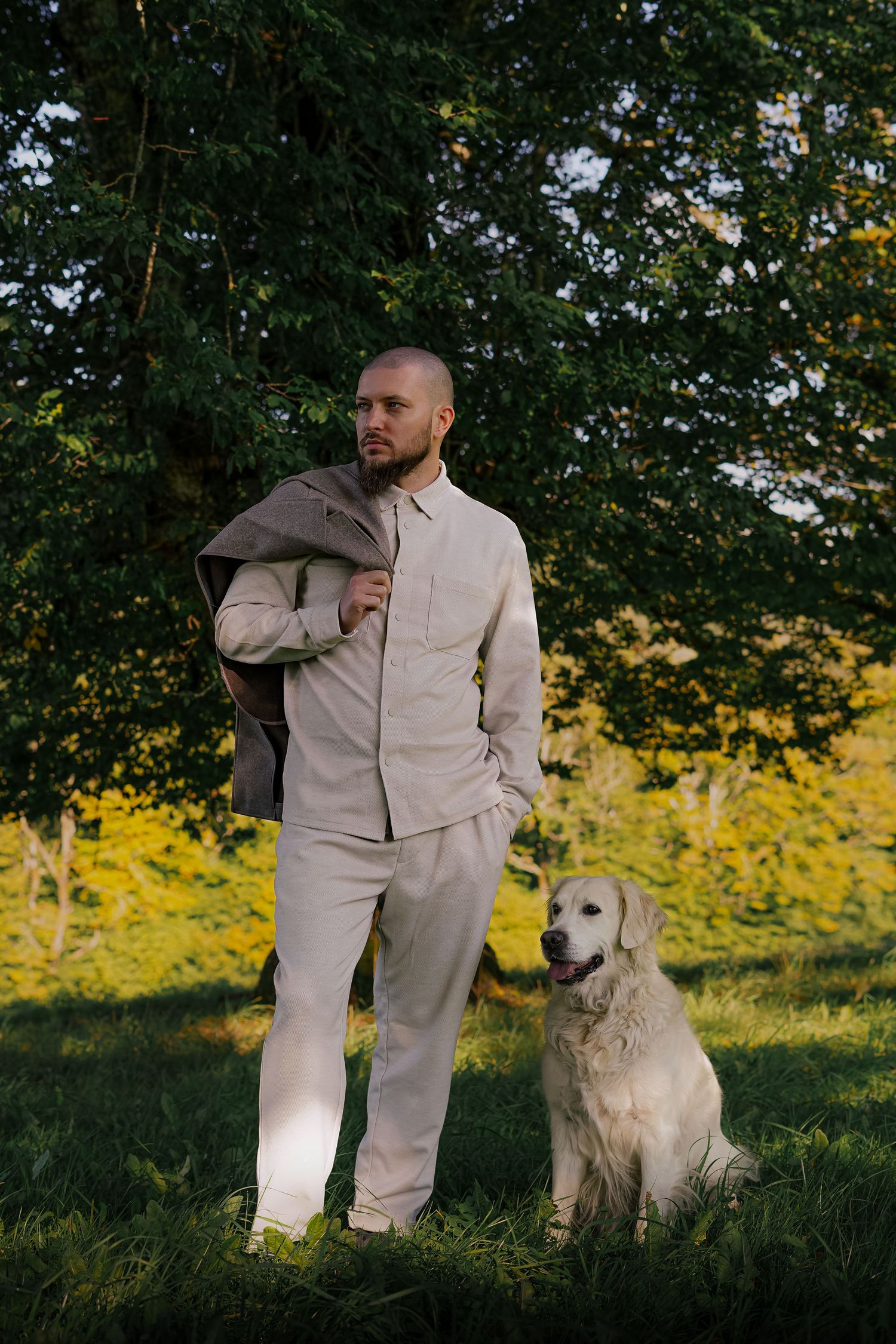 Young man in beige suit standing with golden retriever dog on green grass near trees.