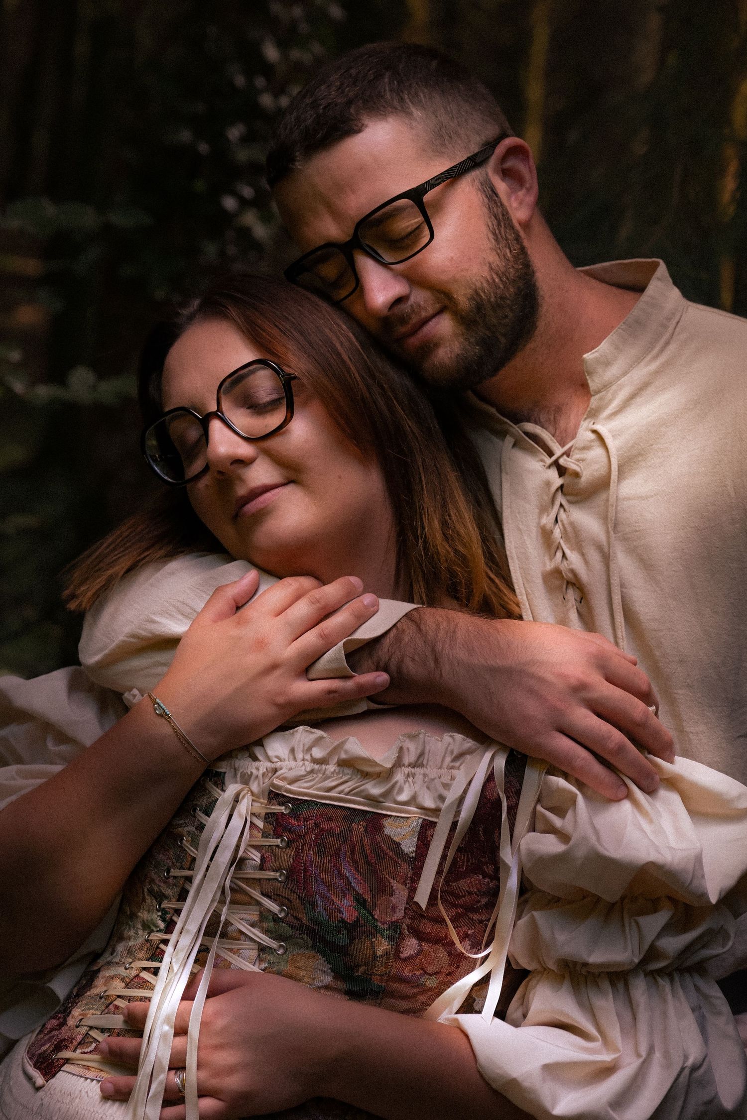 A couple in medieval-style clothing embraces tenderly in a forest, both wearing glasses, eyes closed.