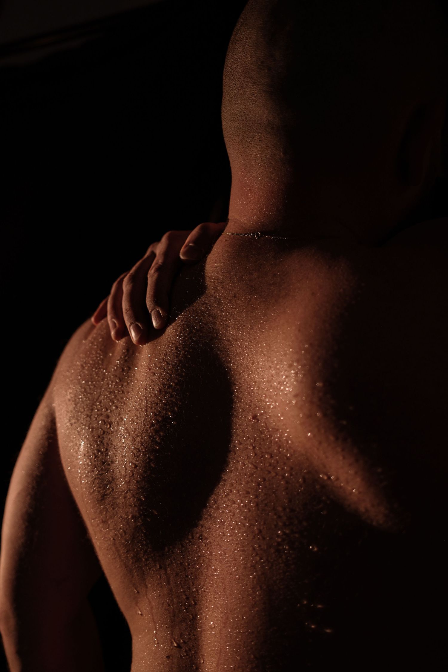 Close-up of muscular back with water droplets and hand on shoulder in dramatic lighting.