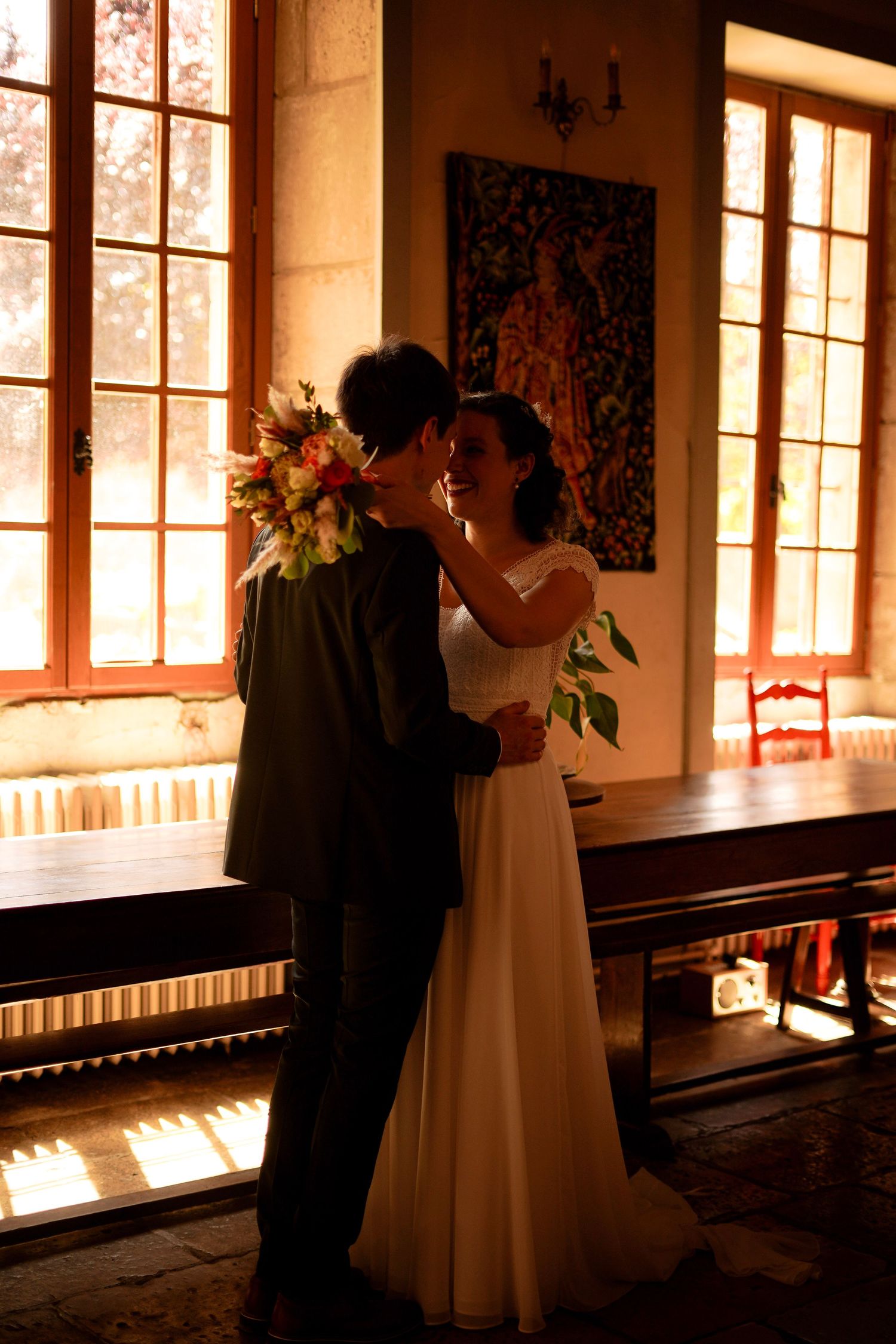 Bride and groom share a romantic moment in a sunlit rustic hall with large windows and a bouquet.