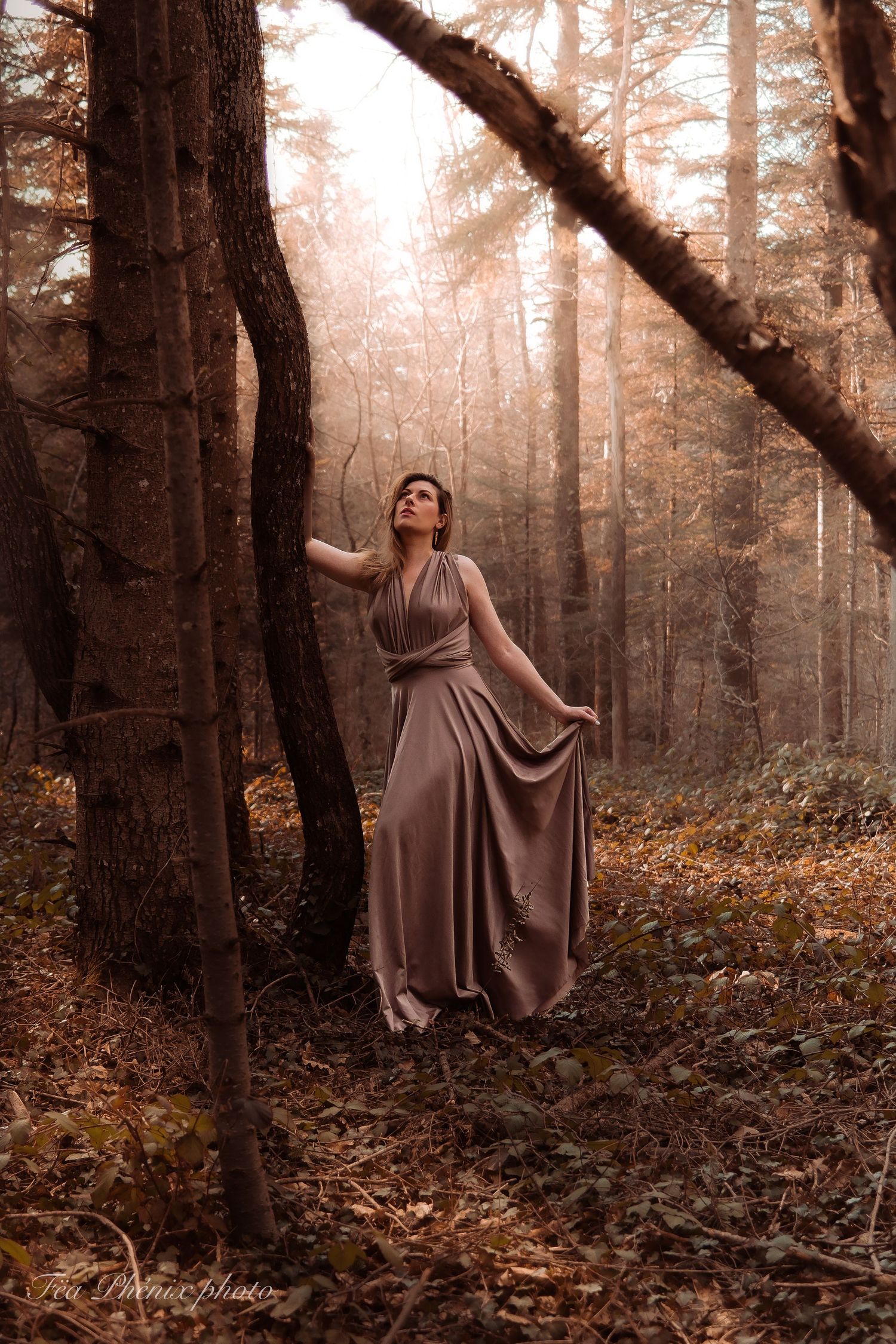 Woman in flowing taupe wrap dress stands between trees in misty autumn forest with golden light rays.