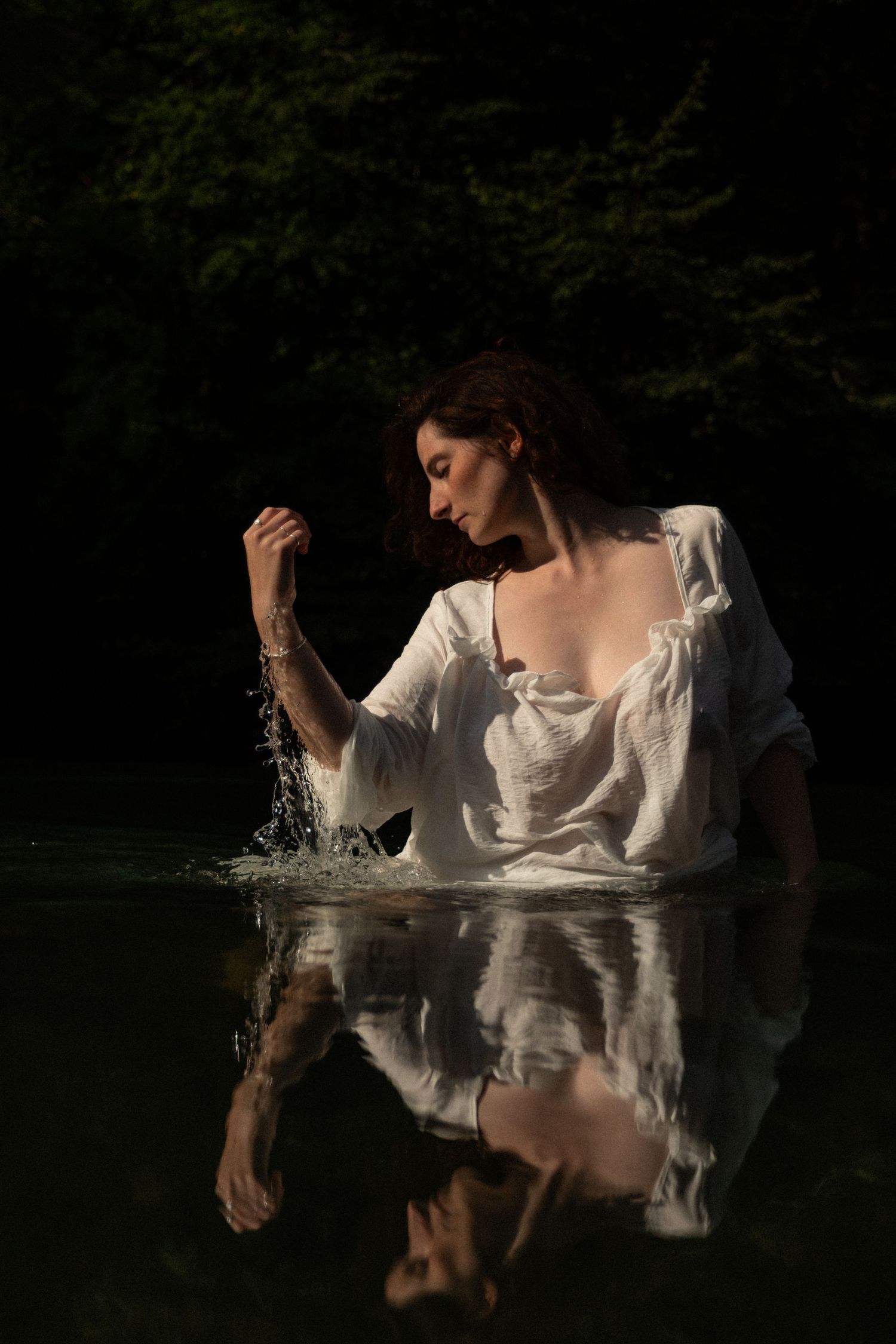 Woman in wet white dress standing in dark forest lake with stunning water reflection.