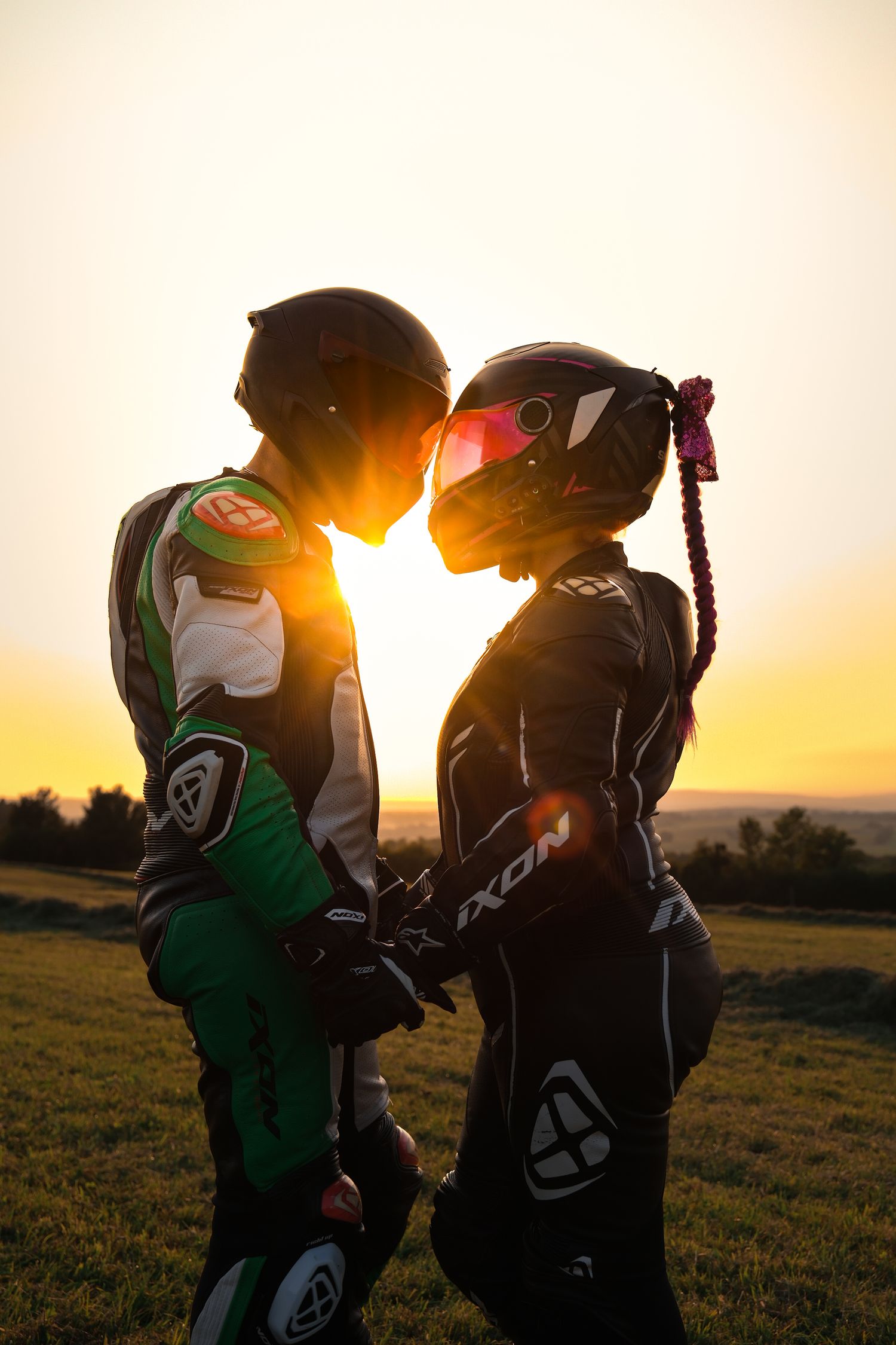 Two motorcyclists in racing gear sharing a romantic moment at golden sunset outdoors.