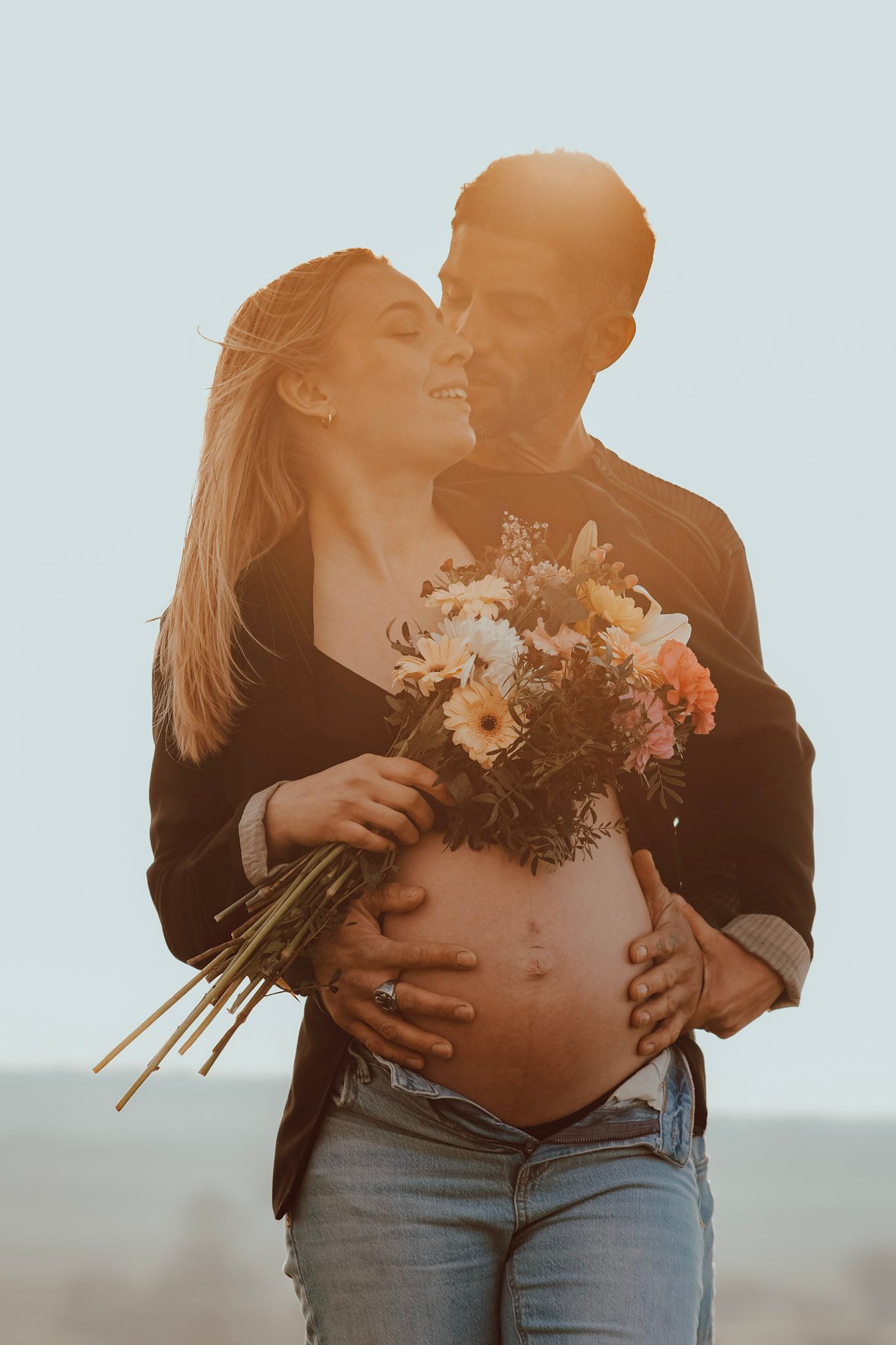 Pregnant couple embracing outdoors at sunset, woman holding wildflower bouquet while partner lovingly cradles her baby bump.