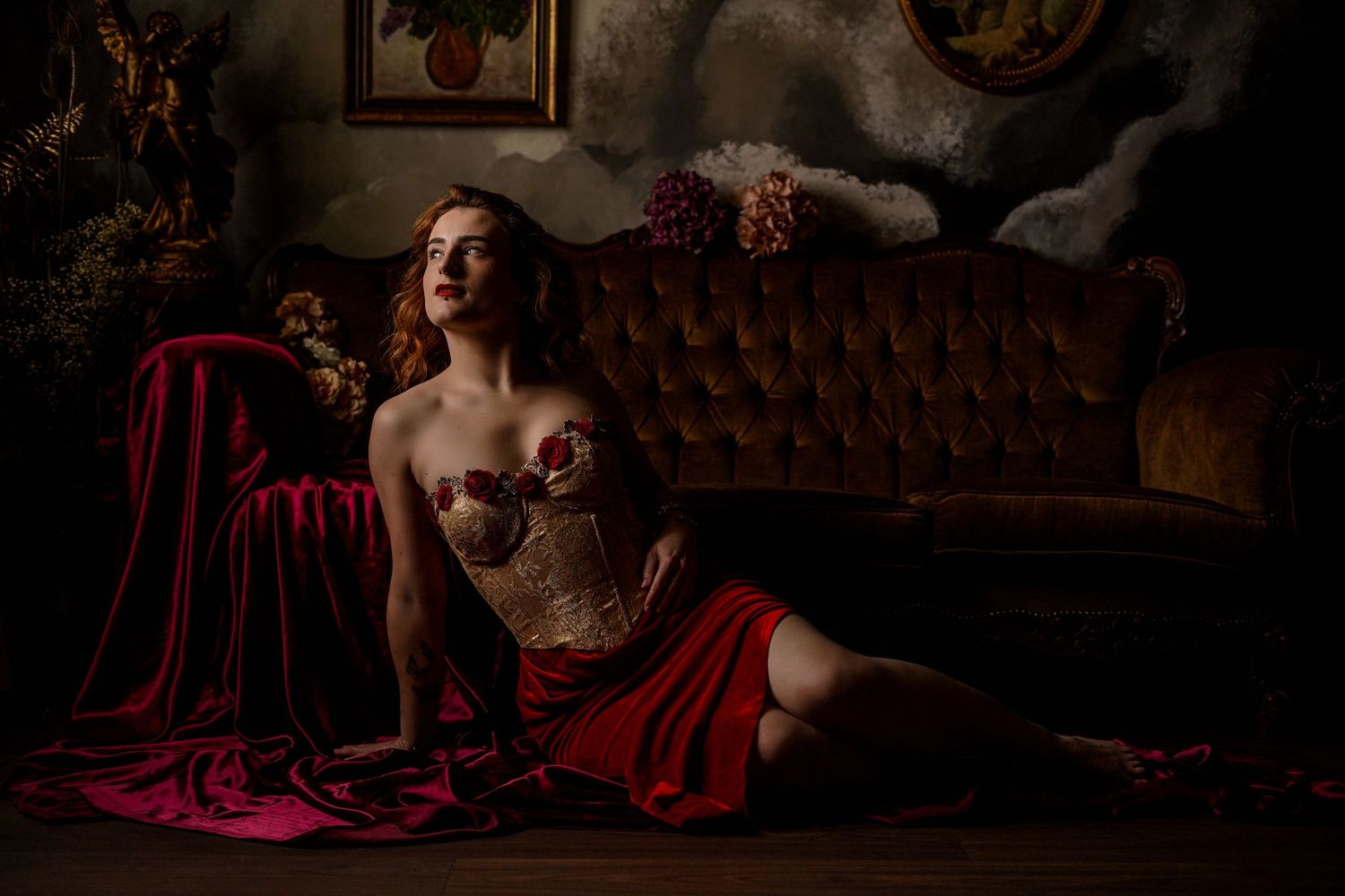 Woman in corset and red skirt poses elegantly on floor beside vintage tufted sofa in dramatic dark studio setting.