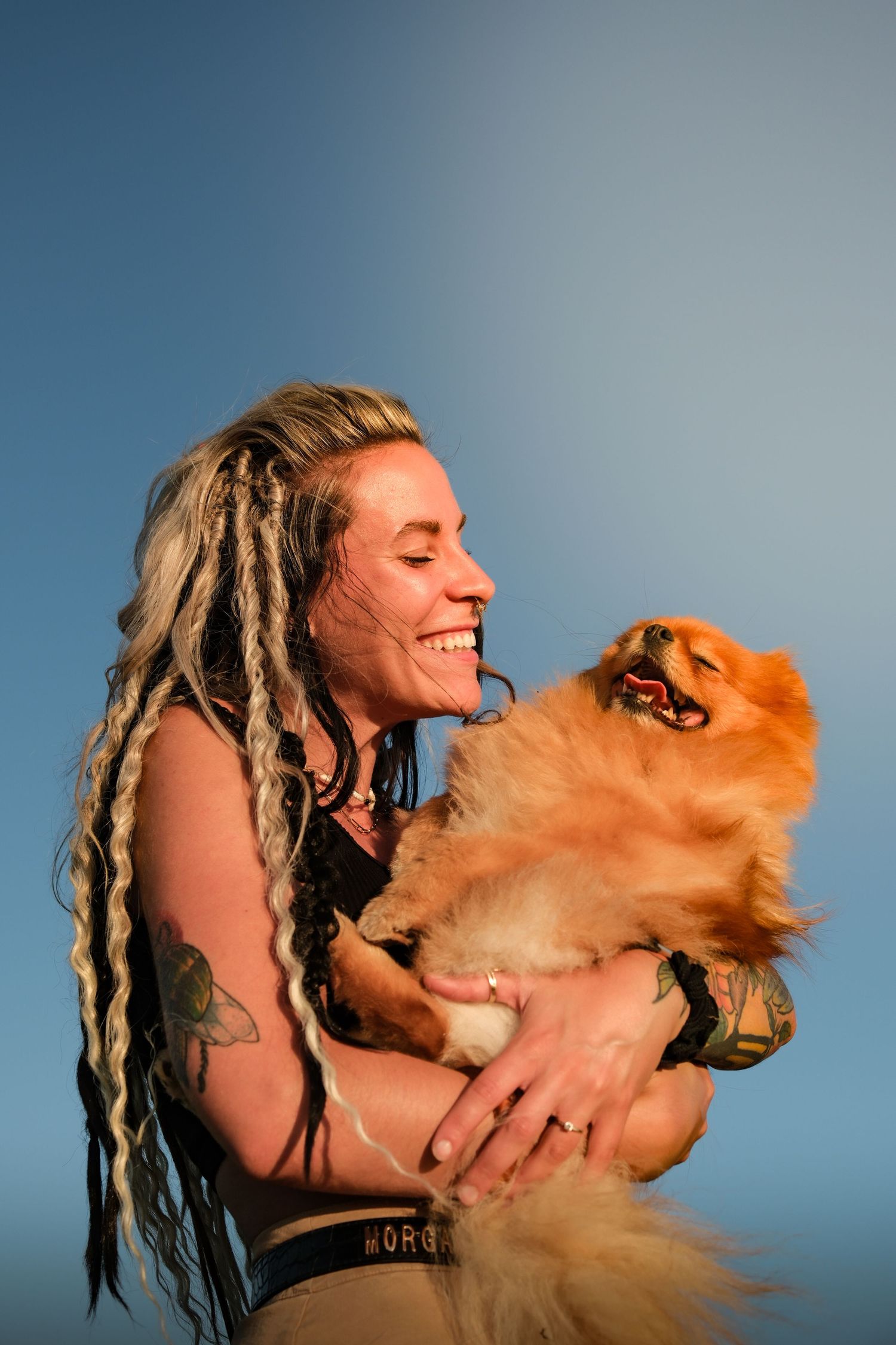 Laughing tattooed woman with dreadlocks holding a fluffy Pomeranian dog against blue sky.