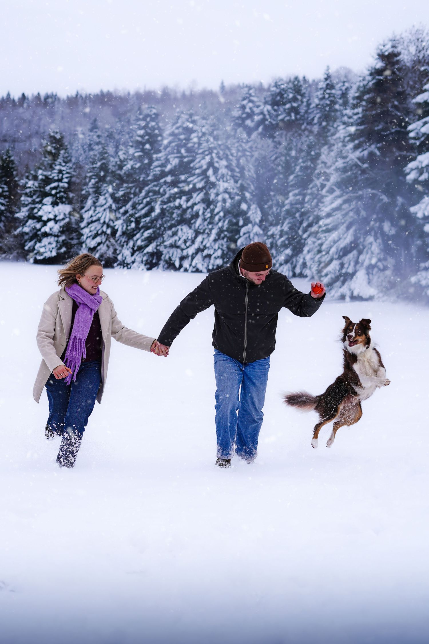 Couple holding hands and running with their Border Collie dog in a snowy winter landscape.