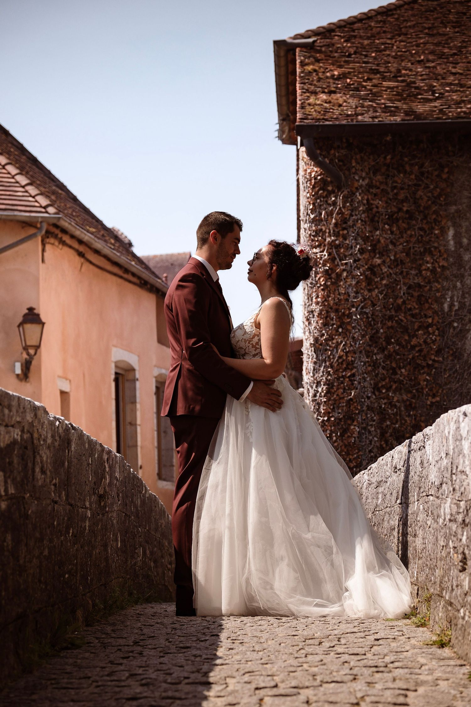 Bride and groom embrace on a cobblestone street between historic buildings under a clear blue sky.