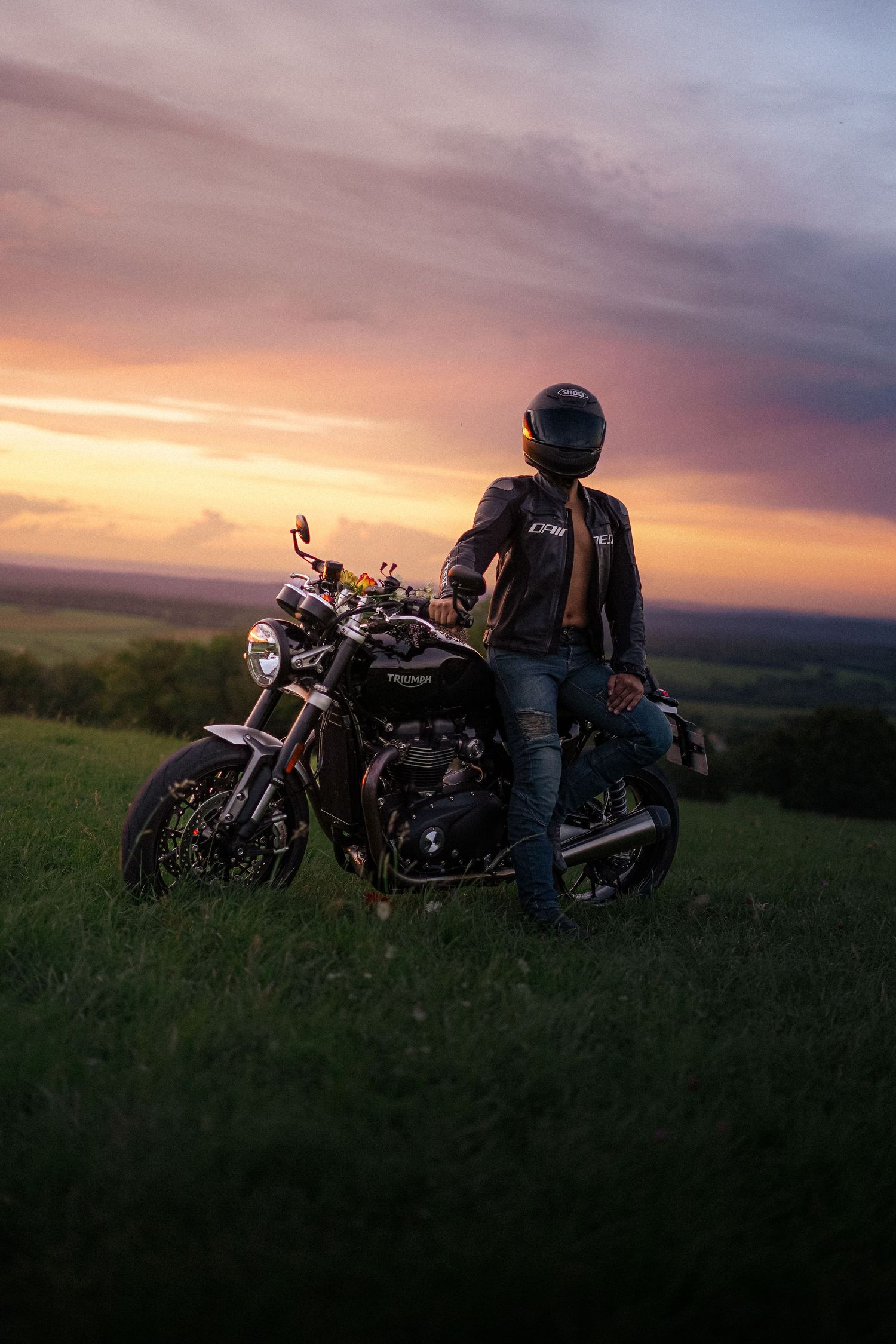 Motorcyclist in leather jacket standing beside Triumph motorcycle at dramatic sunset.