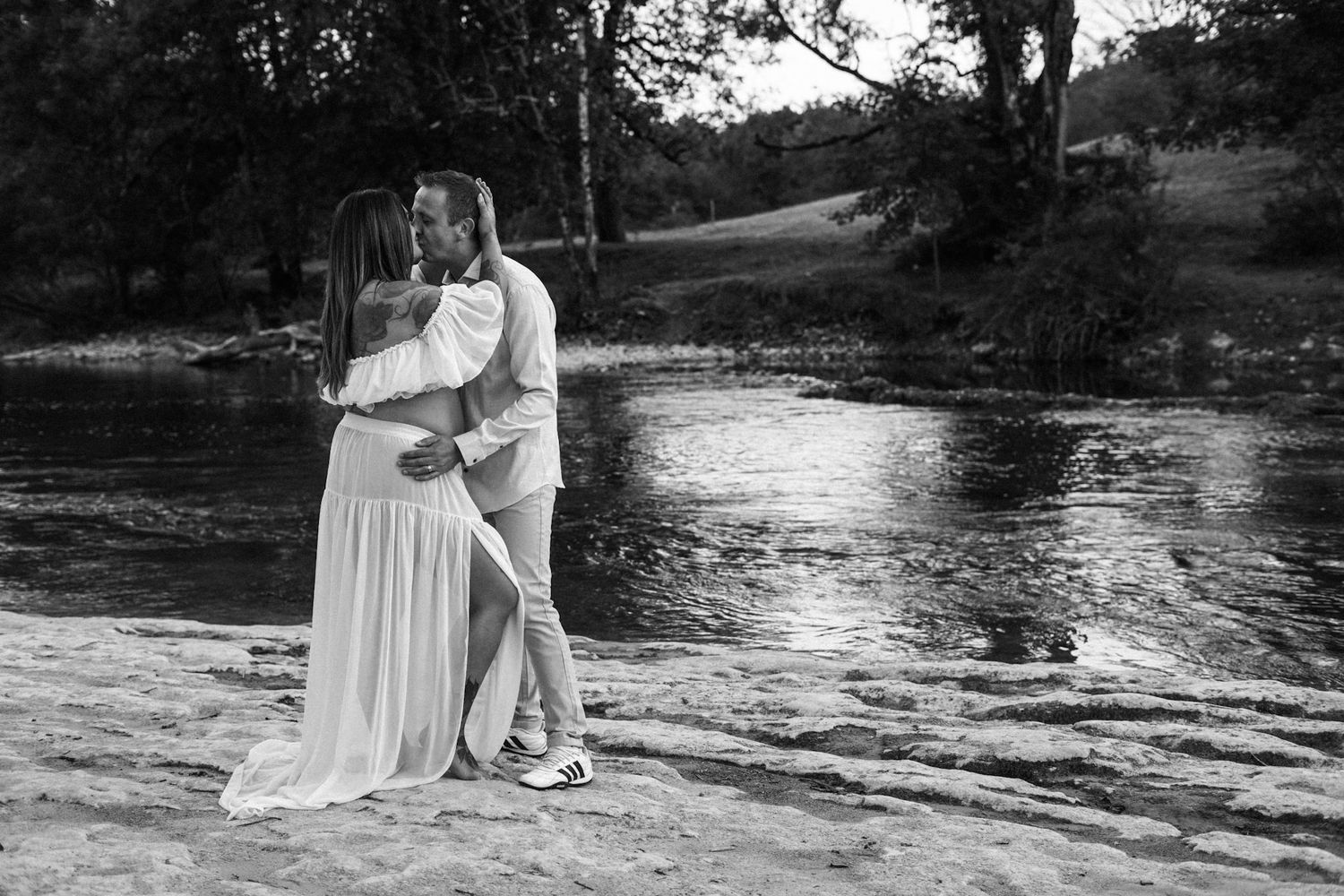 Black and white photo of a couple embracing by a serene river, woman in flowing white dress, romantic outdoor portrait.