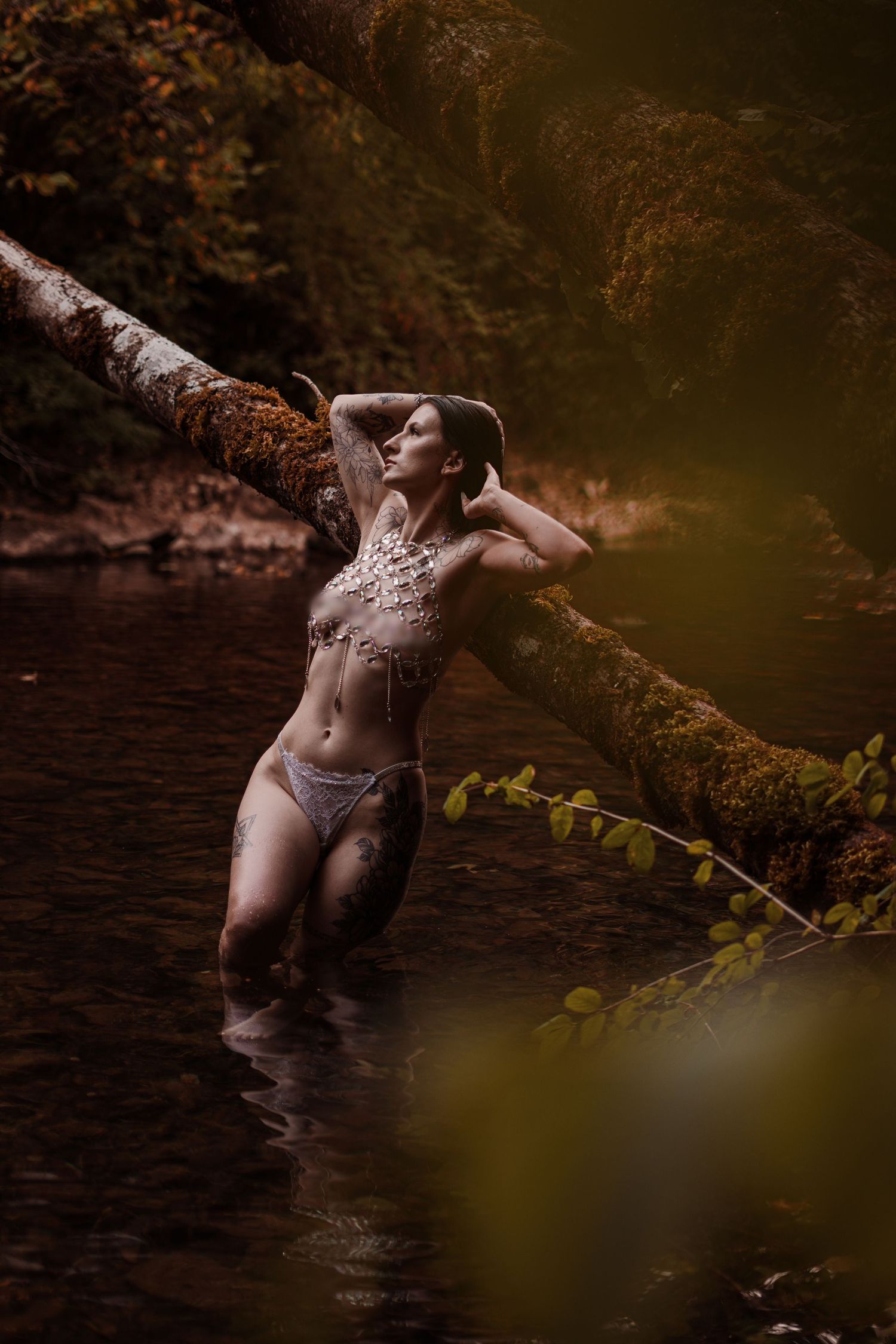 A woman wearing white bikini bottoms and crystal body jewelry stands in a forest stream leaning against a fallen tree.