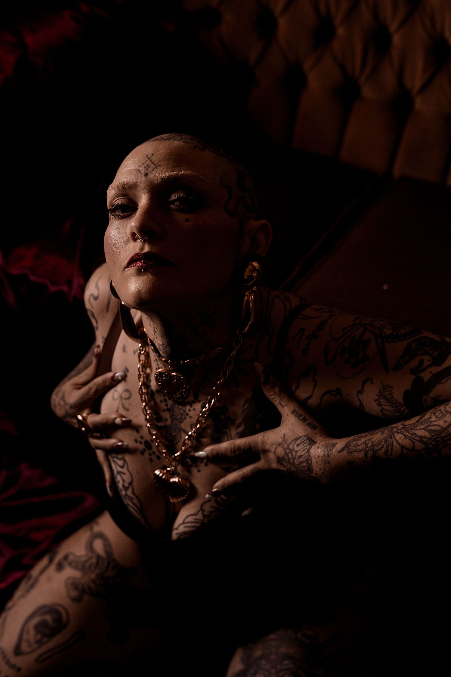 Close-up of tattooed bald woman with gold chain necklace reclining on dark tufted velvet surface.