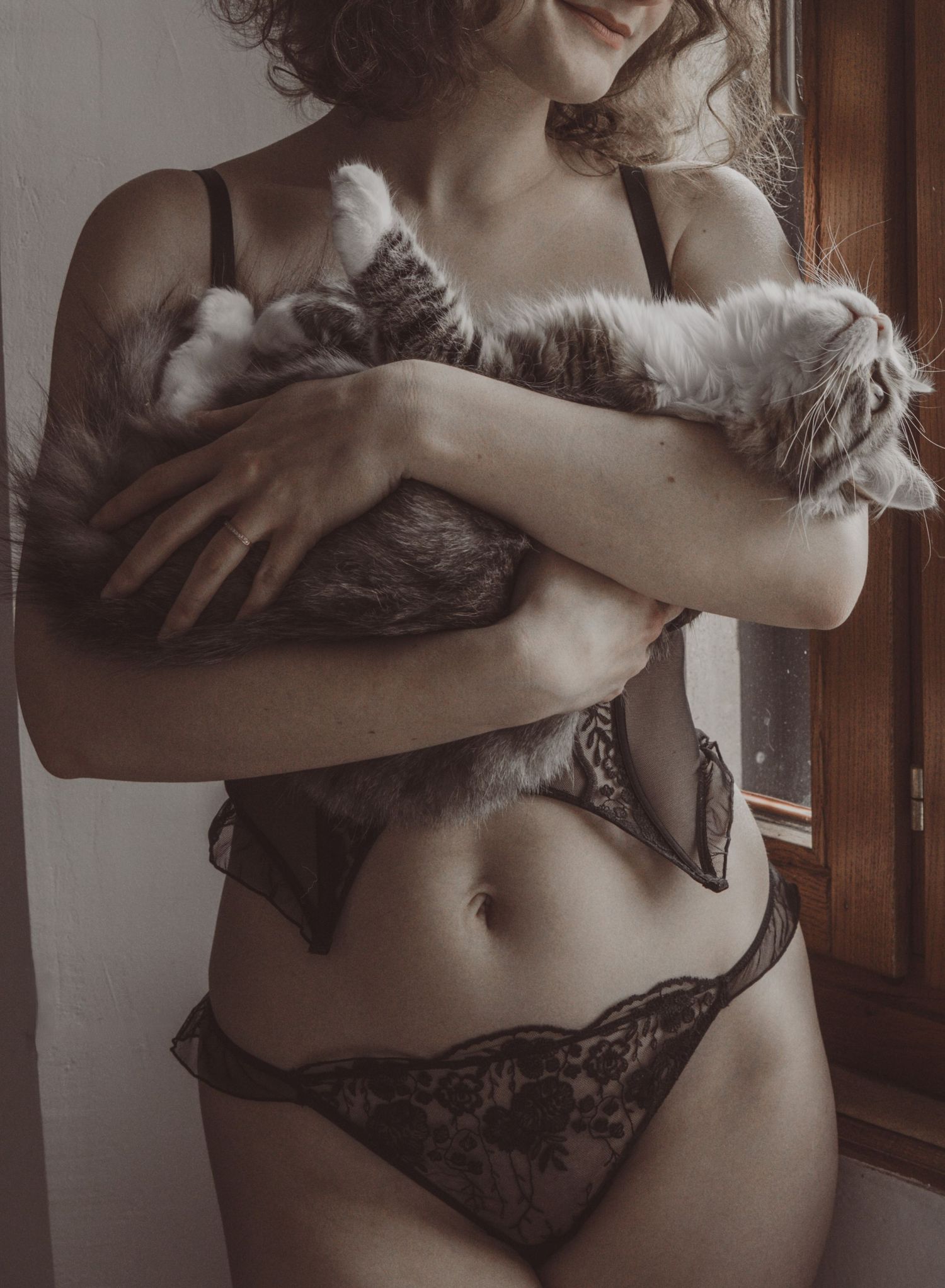 Woman in black lace lingerie holding fluffy gray cat near window.