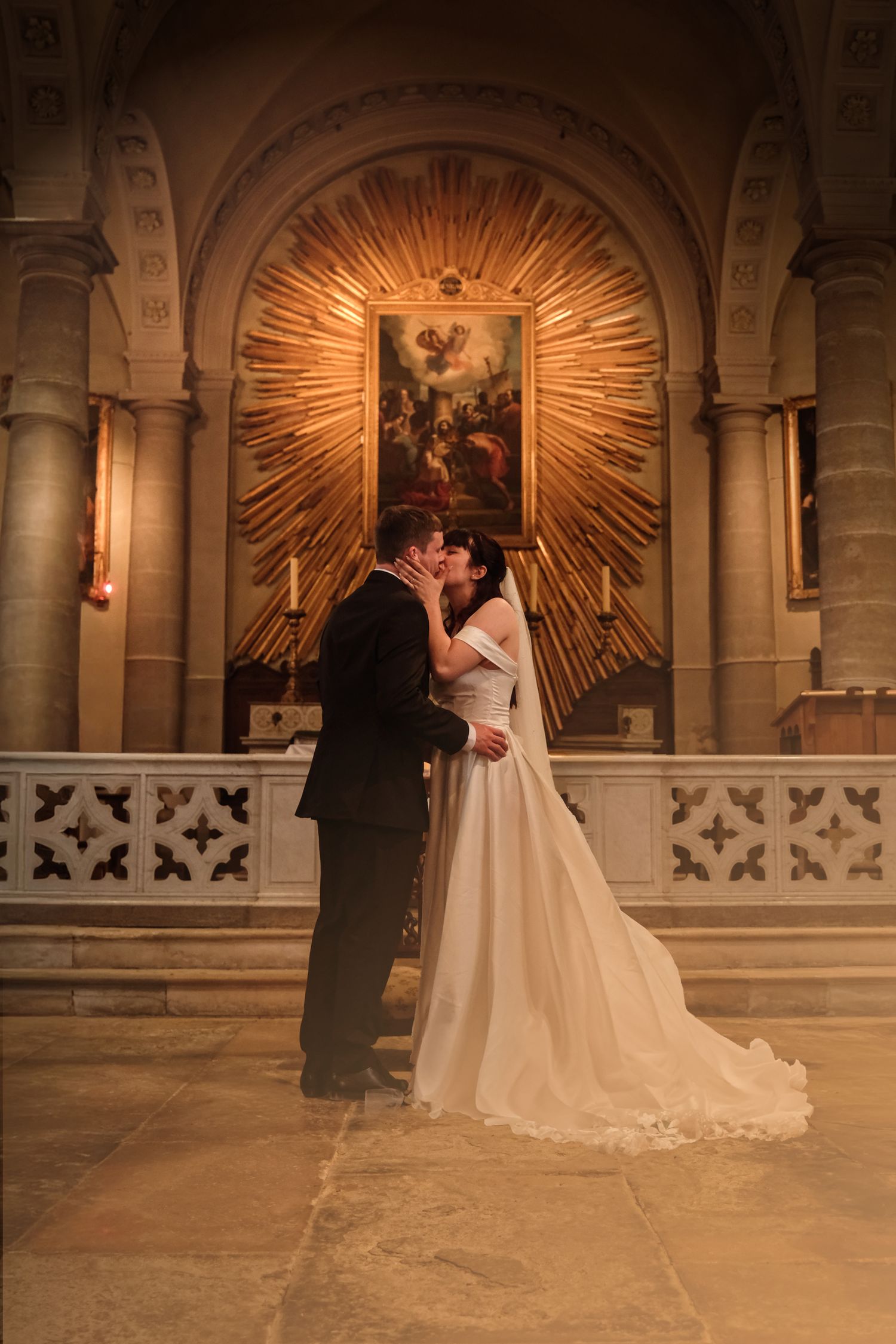 Bride and groom share a romantic first kiss in front of a glowing golden church altar.