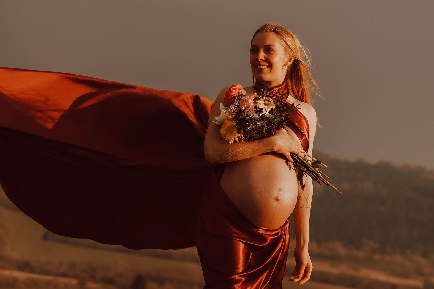 Pregnant woman in flowing rust-red satin dress holding wildflowers in a golden field at sunset.