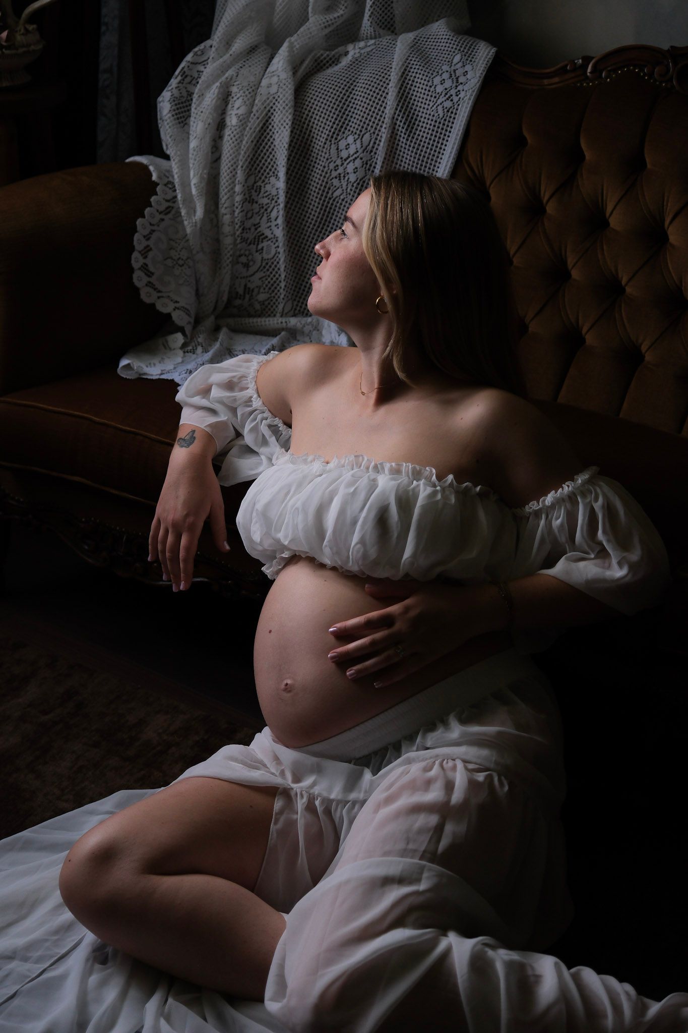 Pregnant woman in white off-shoulder dress sitting on floor leaning against brown velvet sofa in moody dark room.