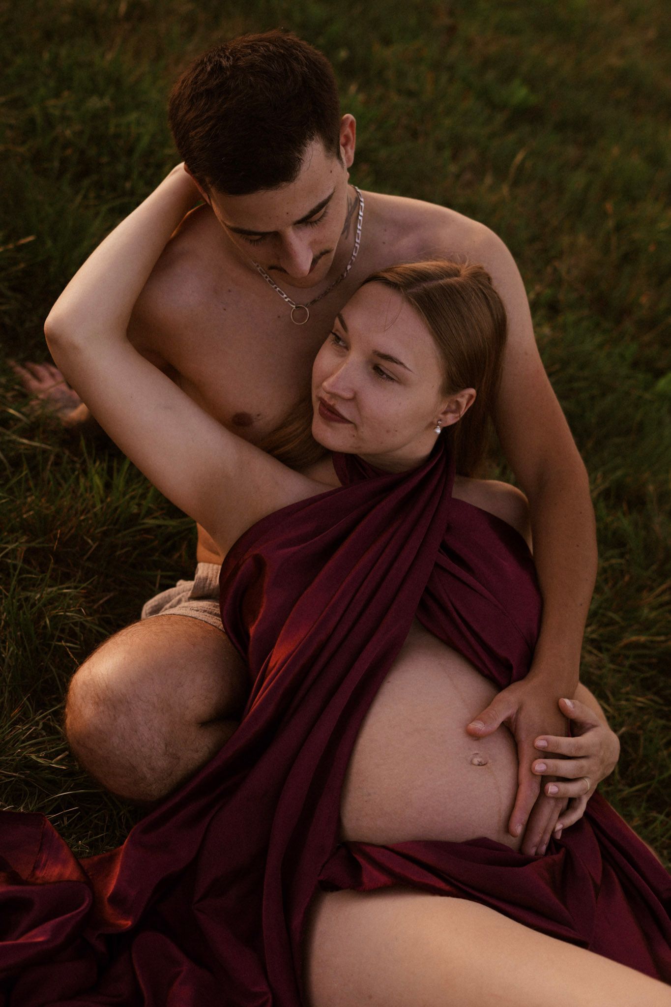 Couple sitting on grass, pregnant woman in burgundy fabric wrap embraced by shirtless man during outdoor maternity photoshoot.