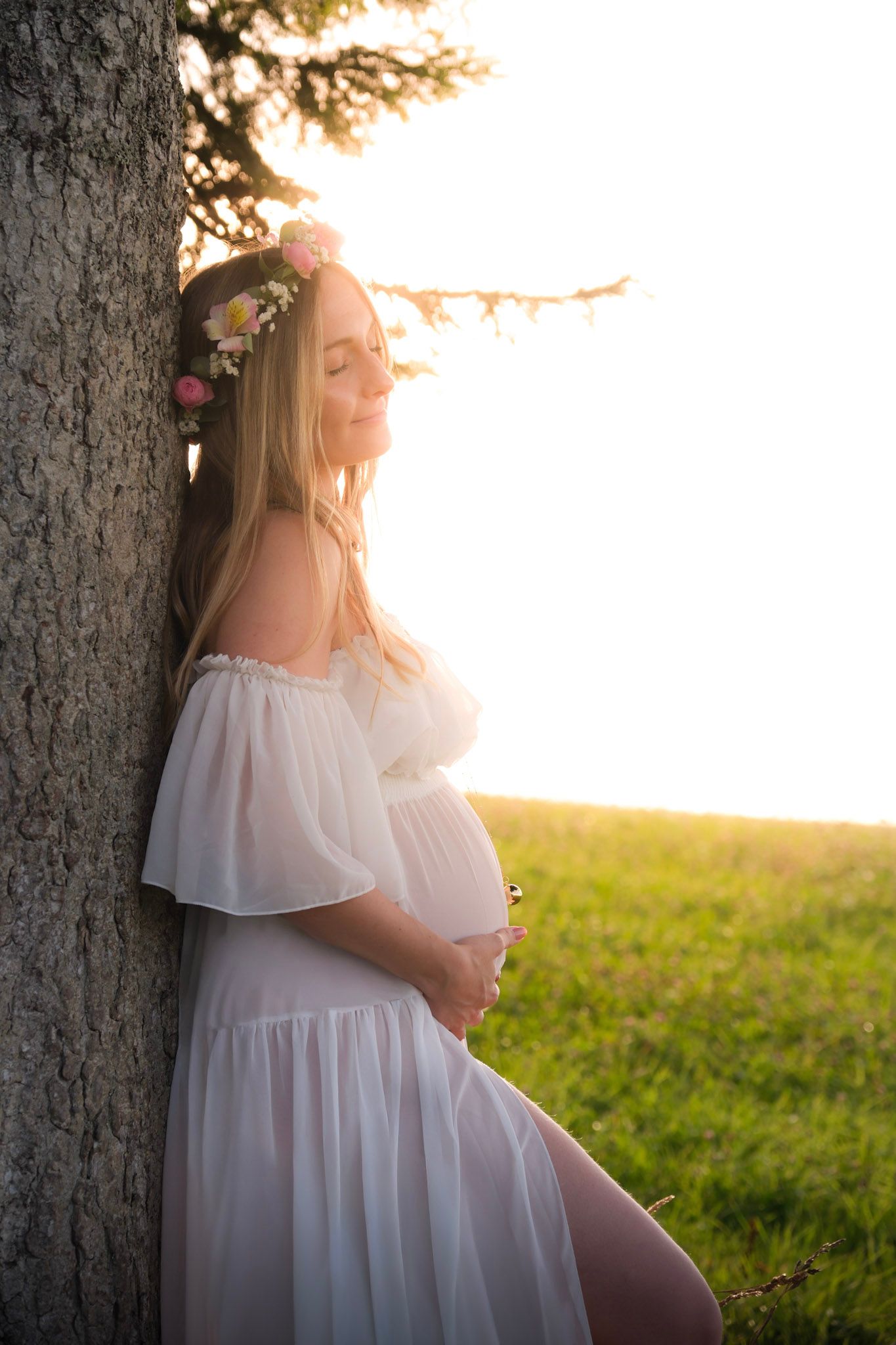 Pregnant woman in white dress and flower crown leaning against tree at golden hour sunset.