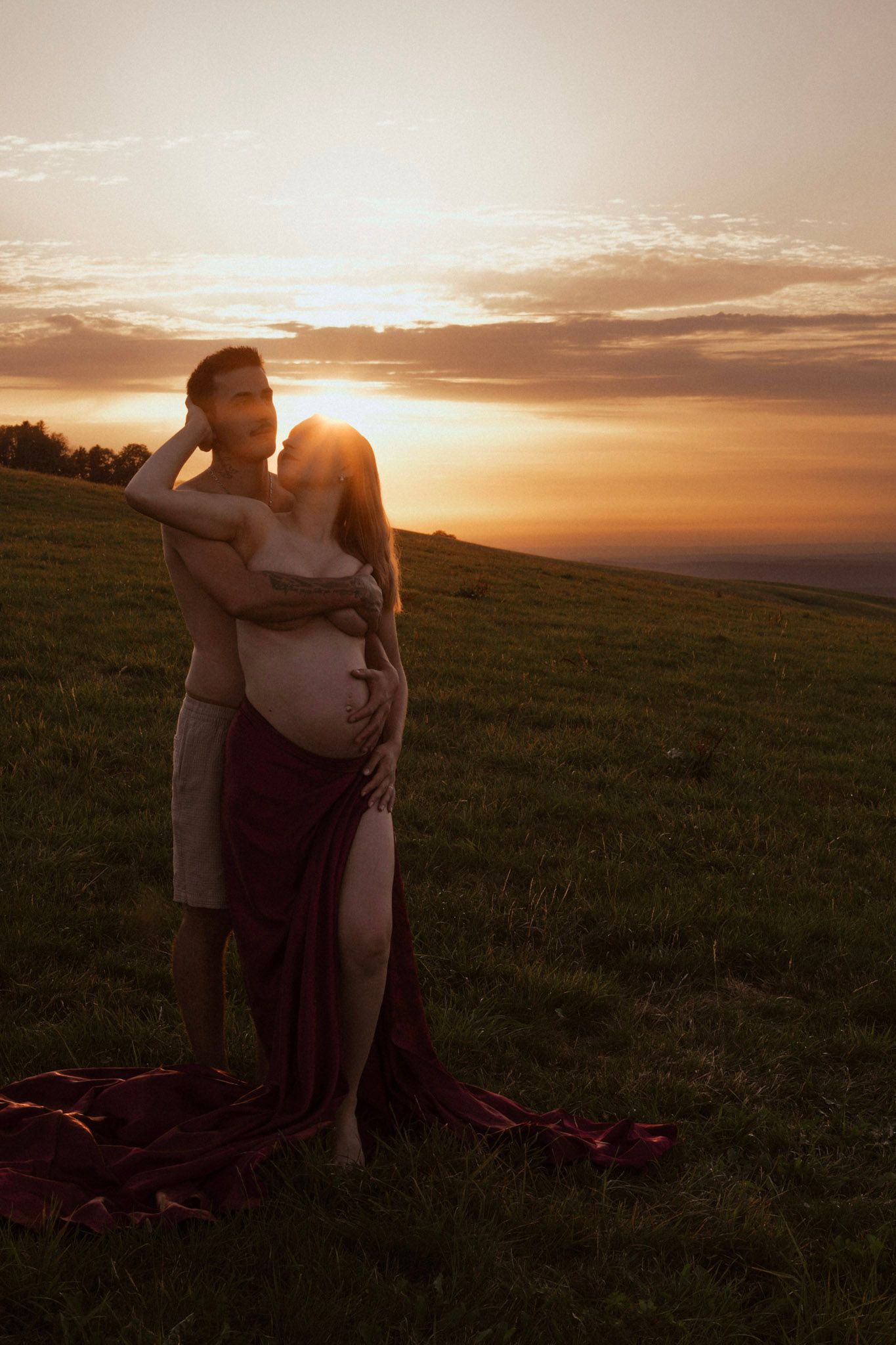 Couple embracing on green hillside at golden sunset, pregnant woman wrapped in red fabric.