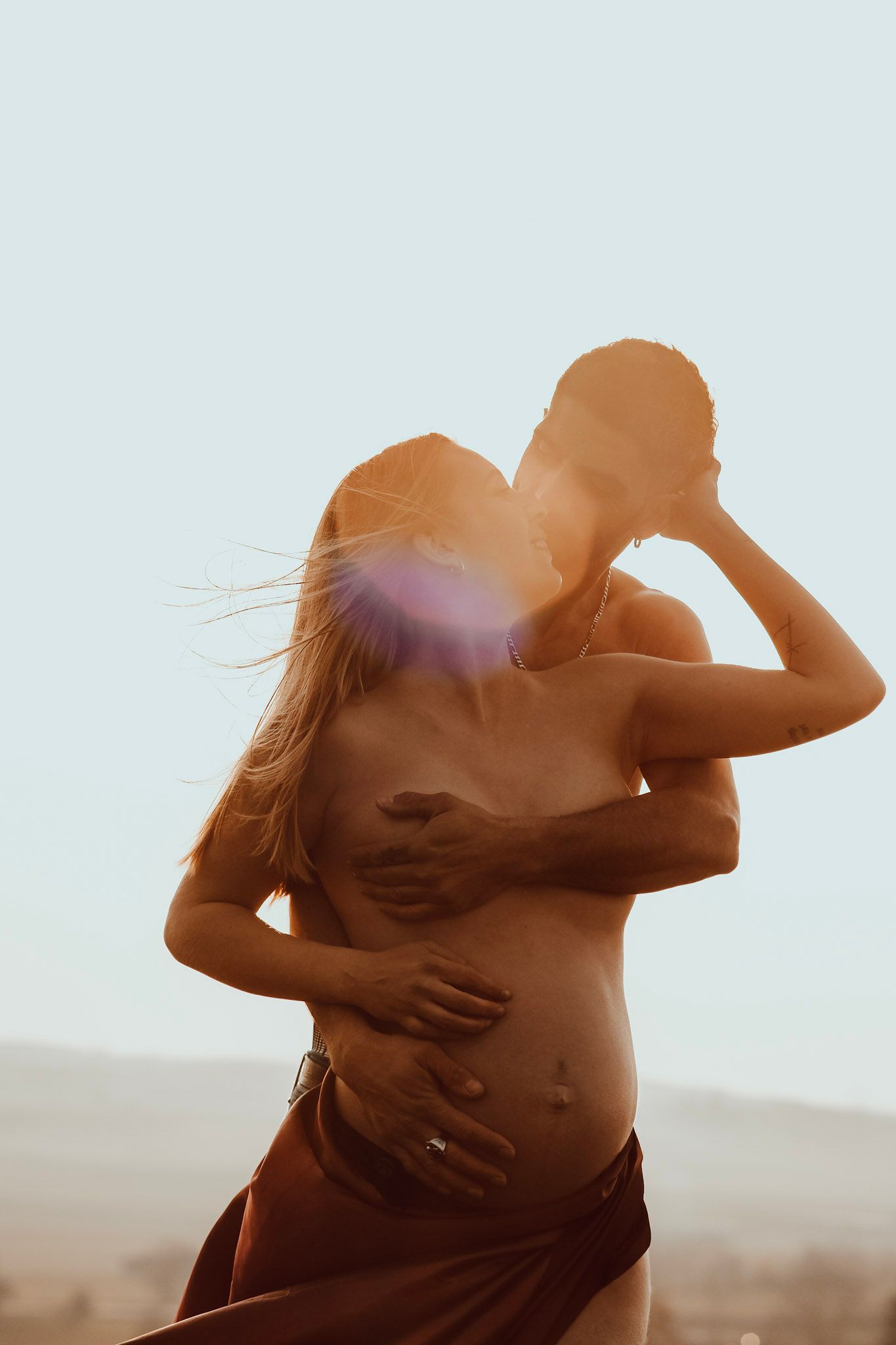 Couple embracing outdoors at golden hour, pregnant woman in brown skirt with wind-blown hair in warm sunlight.