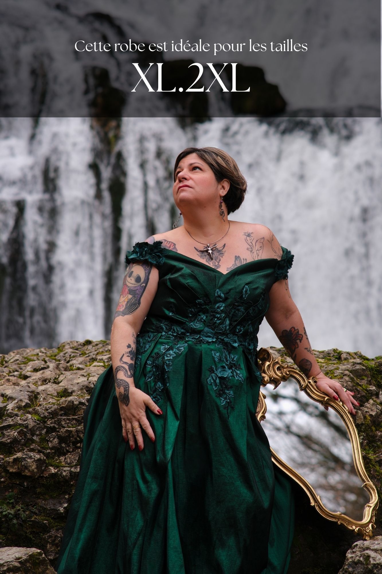Woman in emerald green off-shoulder ball gown with tattoos poses by waterfall, ideal for XL-2XL sizes.
