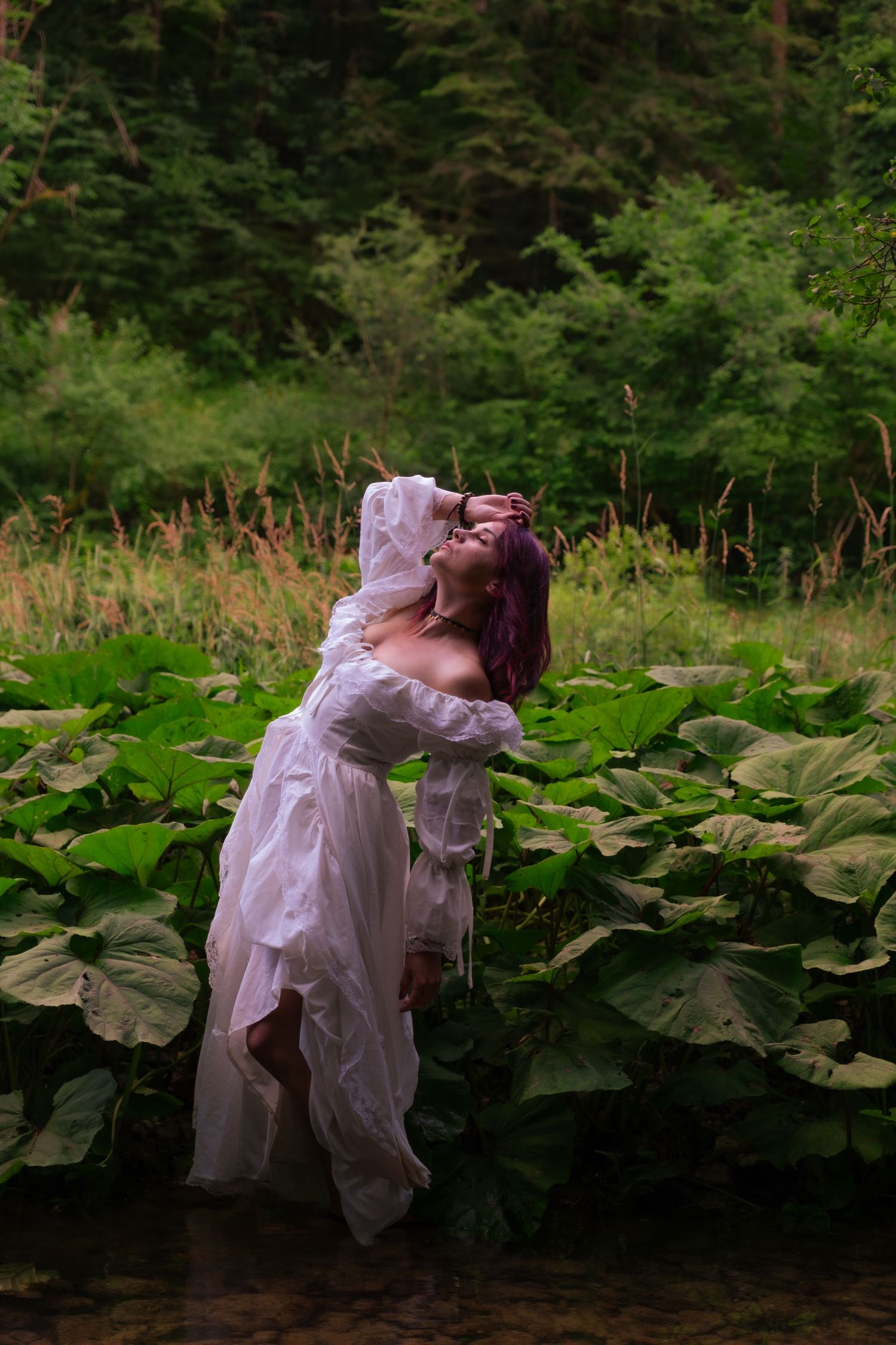 Woman in flowing white dress stands among giant green leaves near water in lush forest setting.