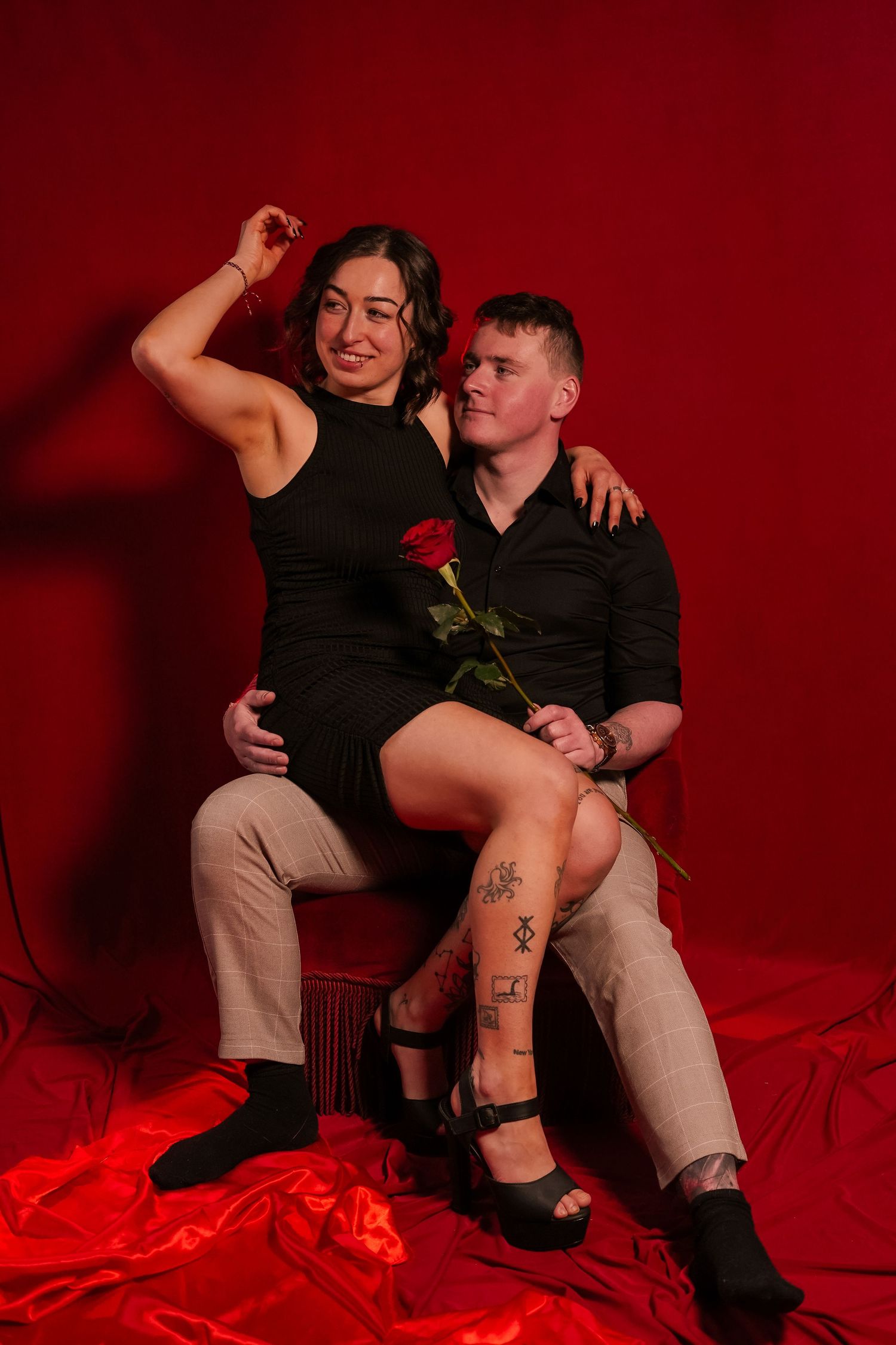 Smiling couple in red studio, woman in black dress sitting on mans lap holding red rose.