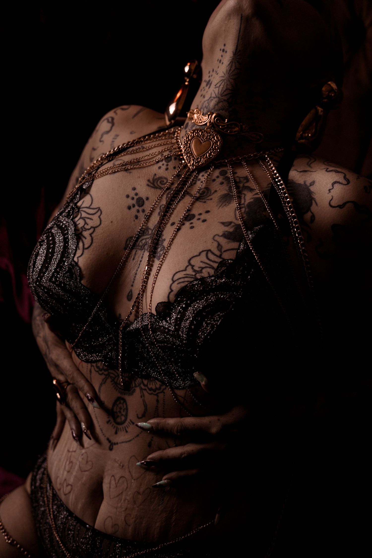 Woman in black lace bra with extensive tattoos and gold chain jewelry in dramatic dark lighting.