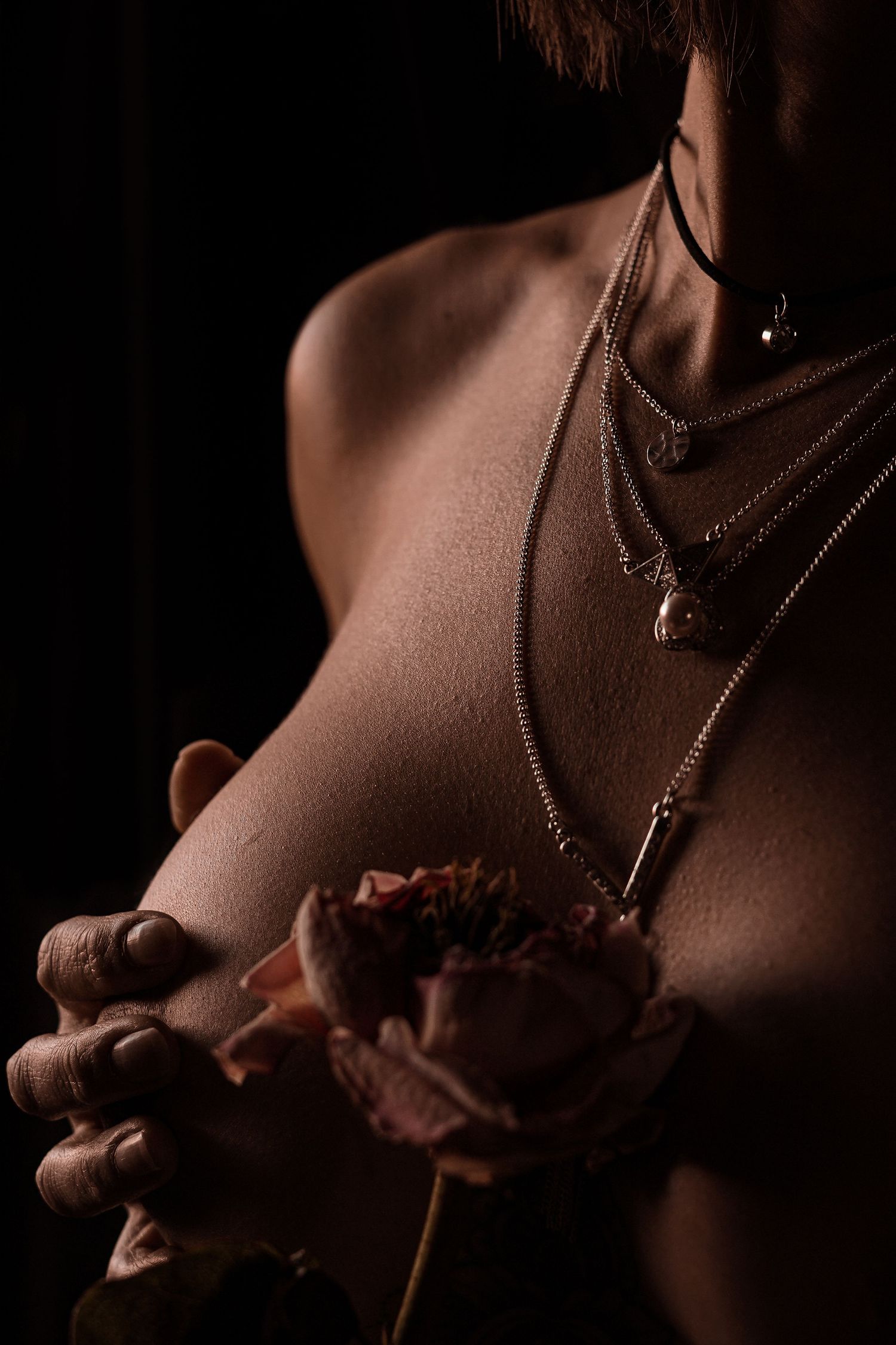 Shirtless person wearing layered necklaces holding a pink rose in moody lighting.
