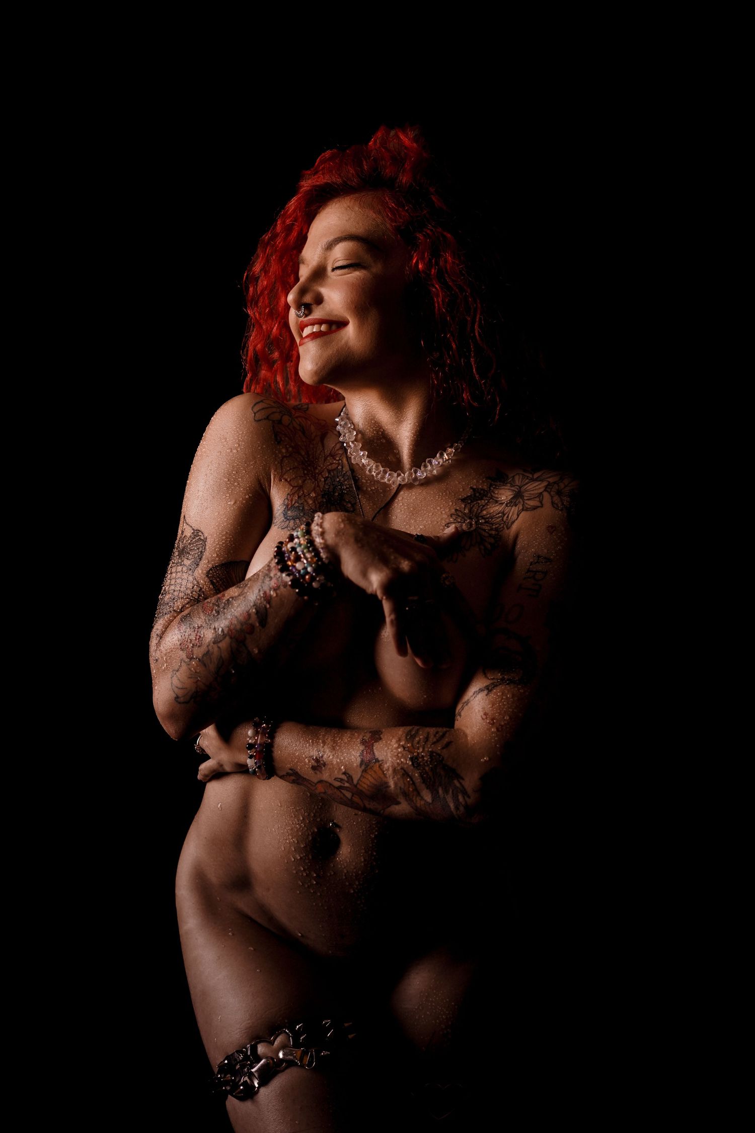 Smiling red-haired tattooed woman posing topless with pearl necklace on black background.