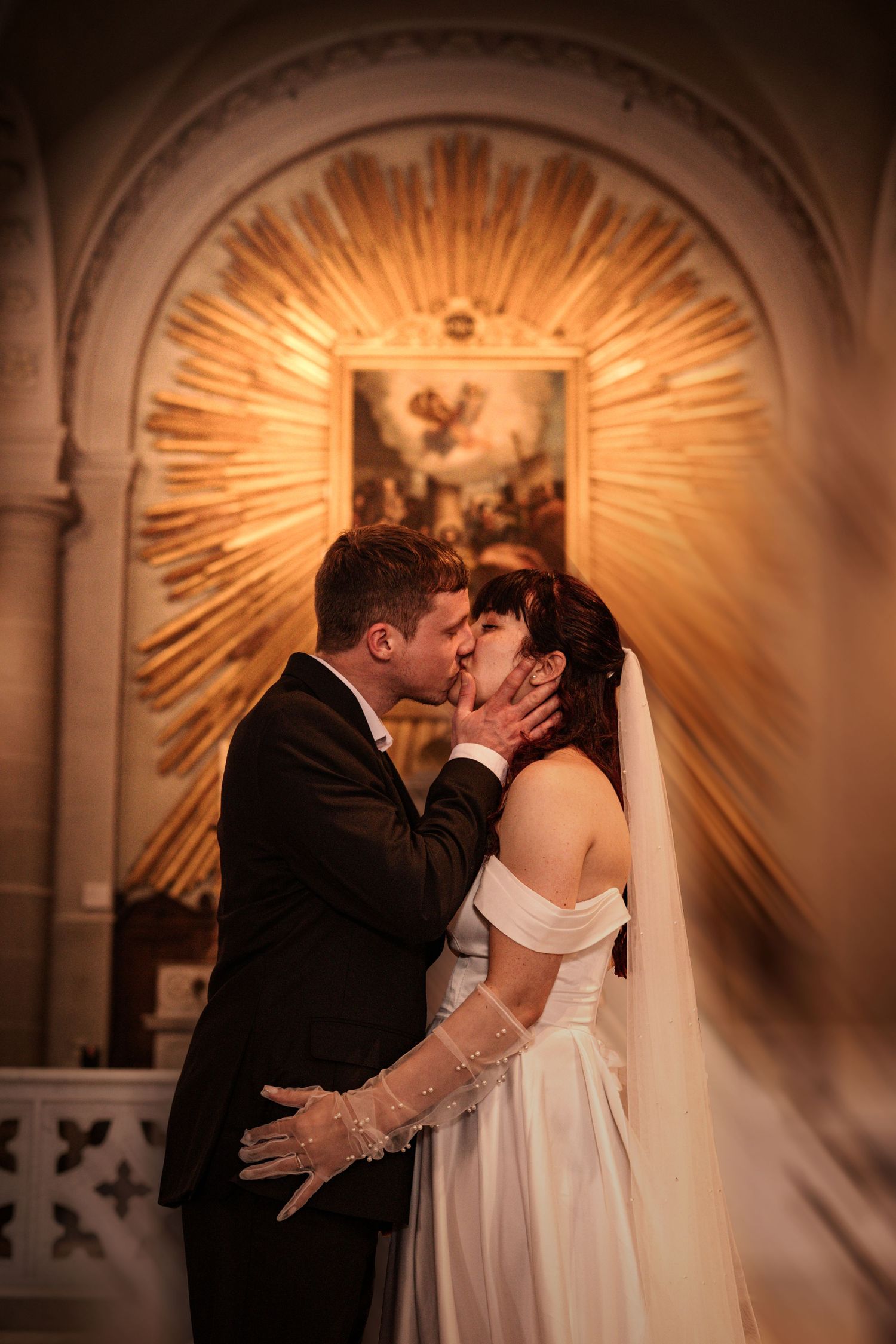 Newlyweds kiss passionately in front of a radiant golden sunburst altarpiece inside a church.