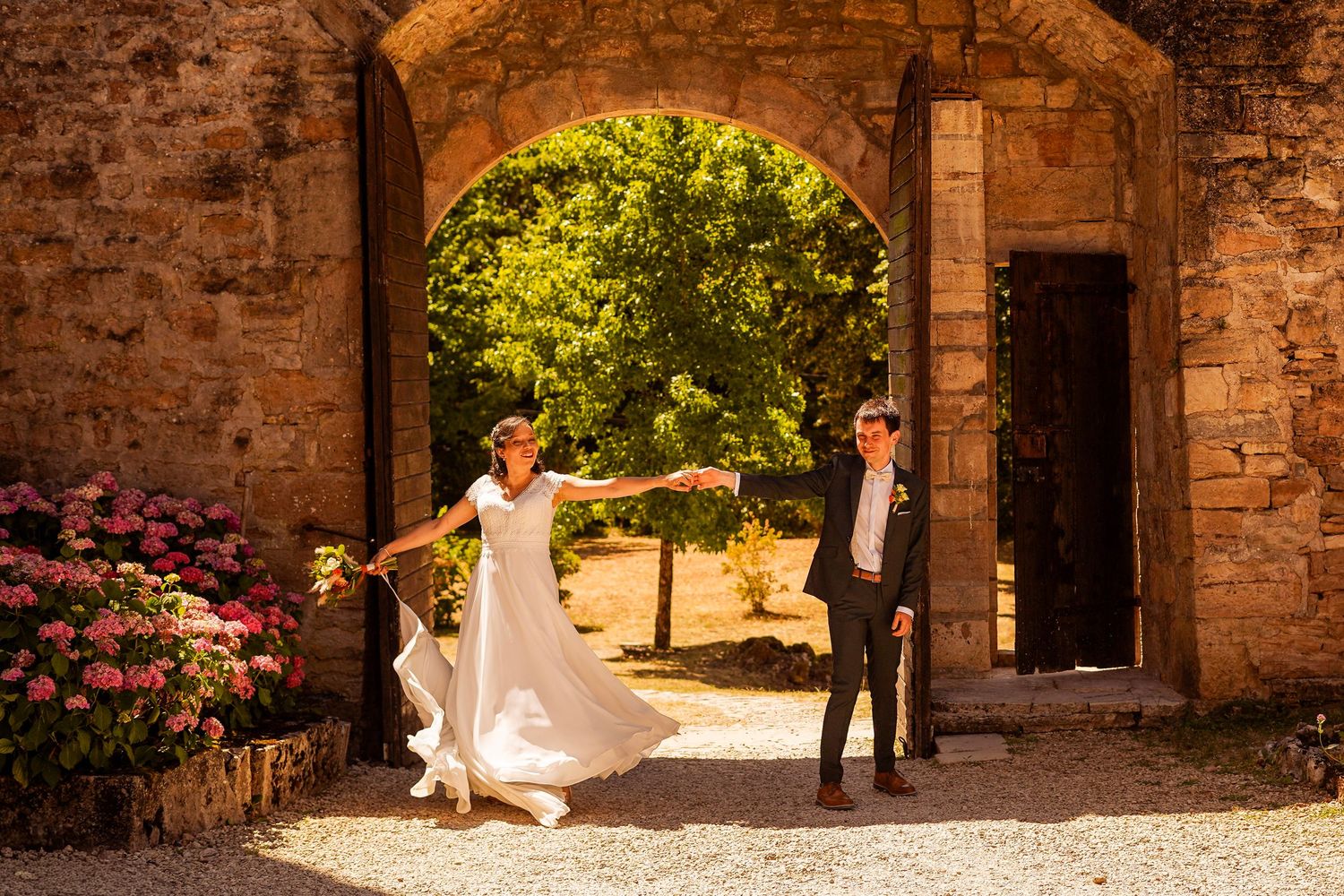 Bride and groom pose playfully under a stone archway with pink flowers at a French chateau venue.