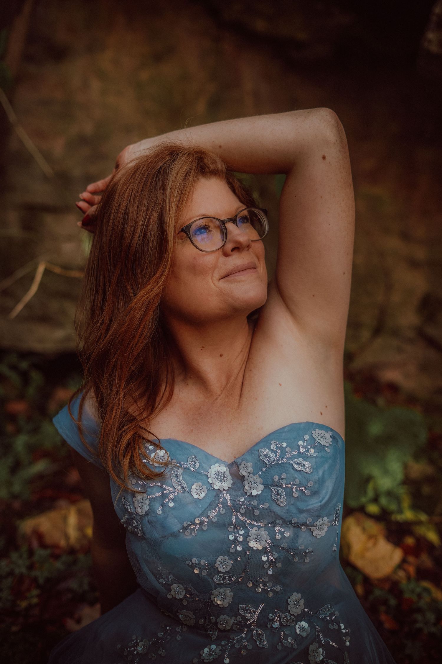 Redhead woman in blue floral strapless gown posing outdoors in forest with arms raised.