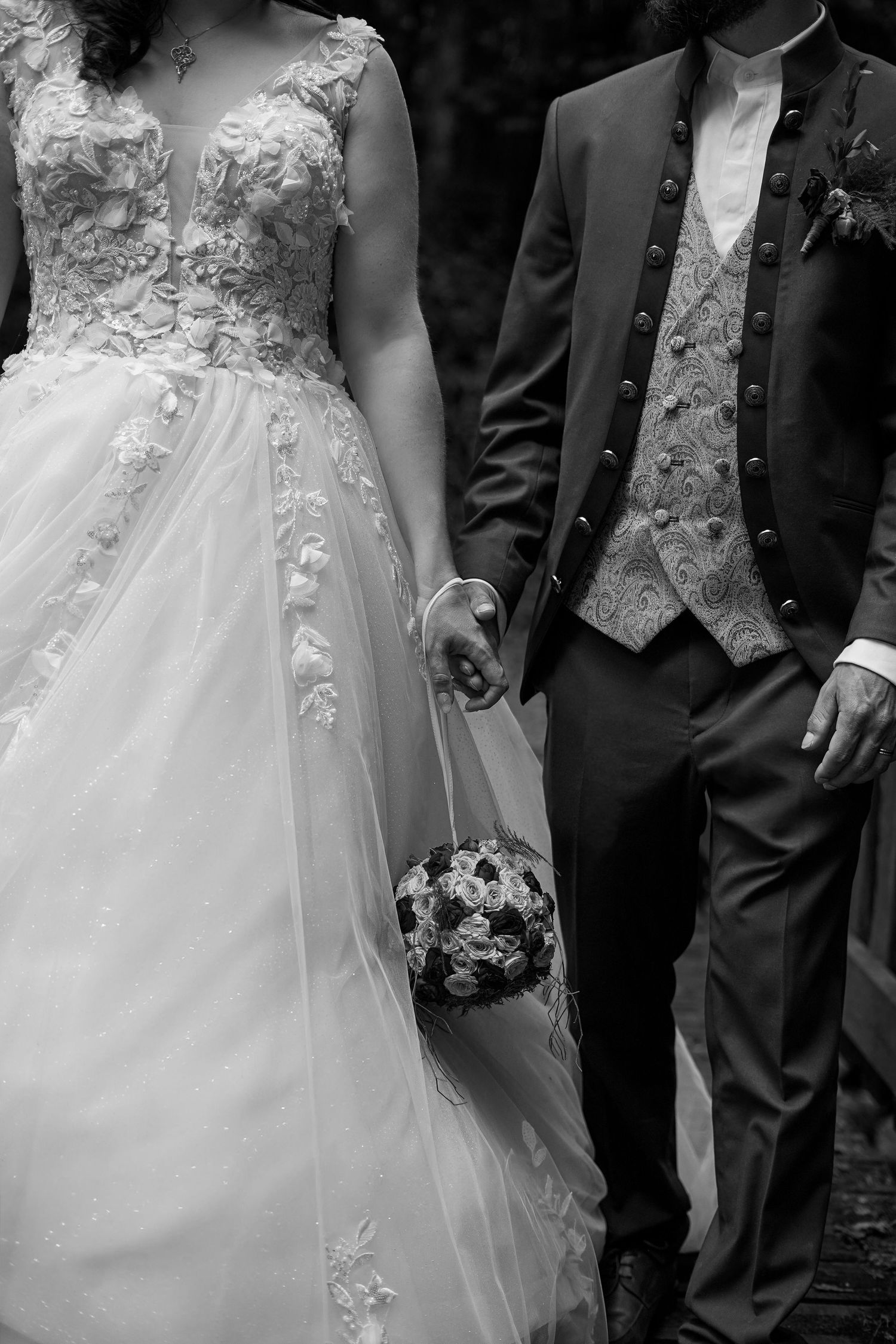 Black and white photo of bride in lace gown and groom in traditional suit holding a bouquet.