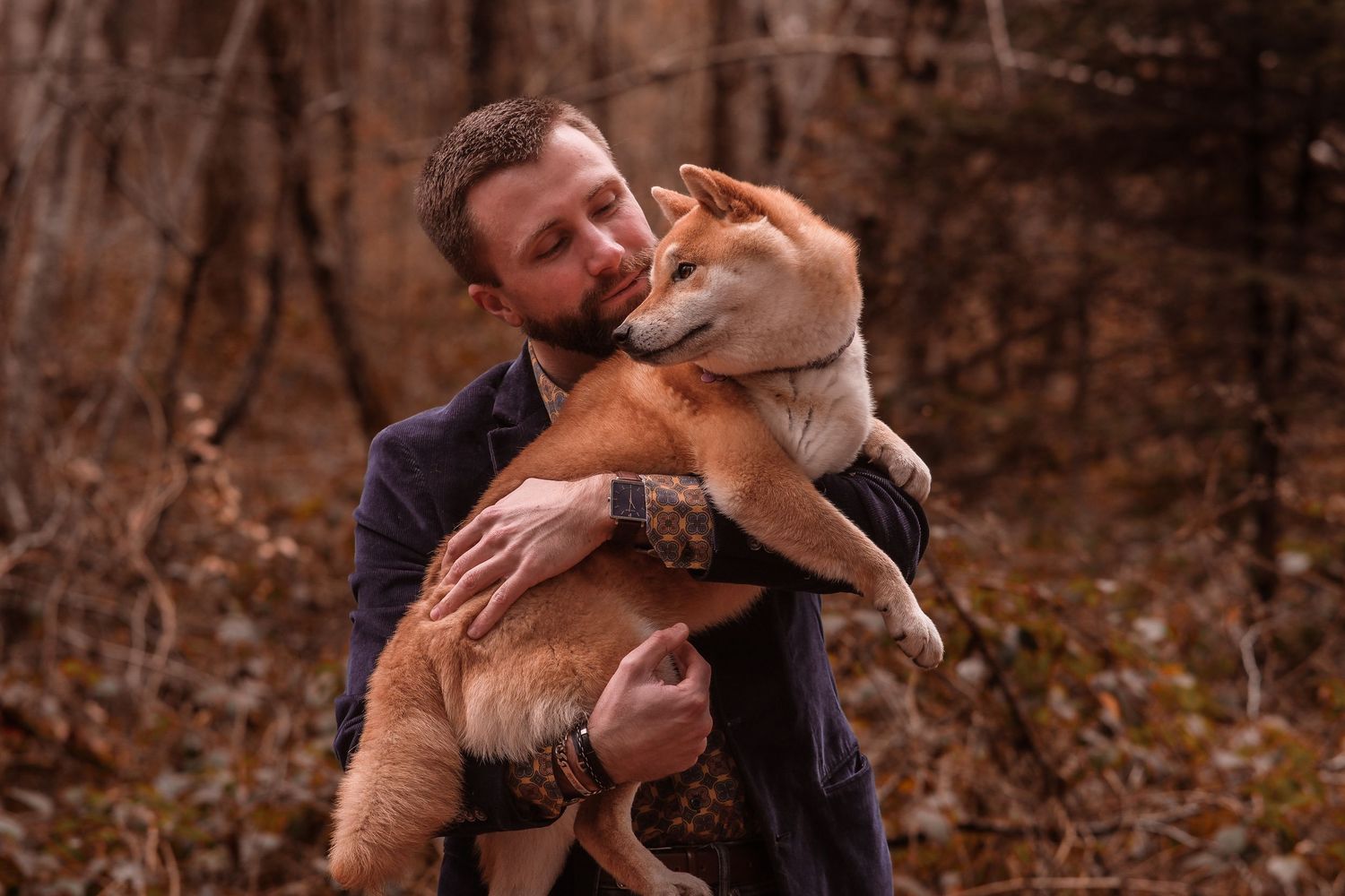 A man holding and cuddling a Shiba Inu dog in an autumn forest.