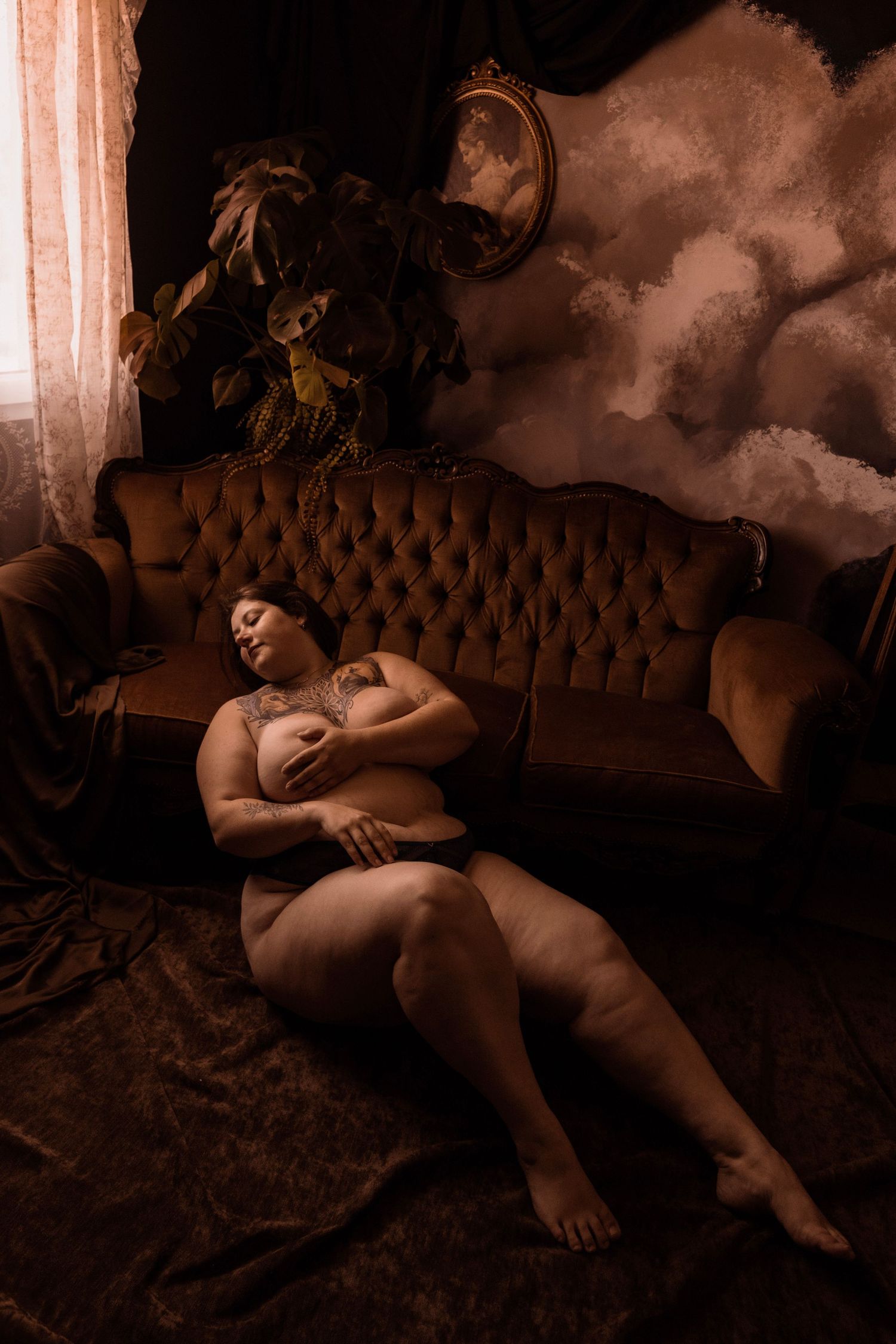 Tattooed woman in lingerie reclining on floor beside vintage velvet sofa in dramatic dark room.