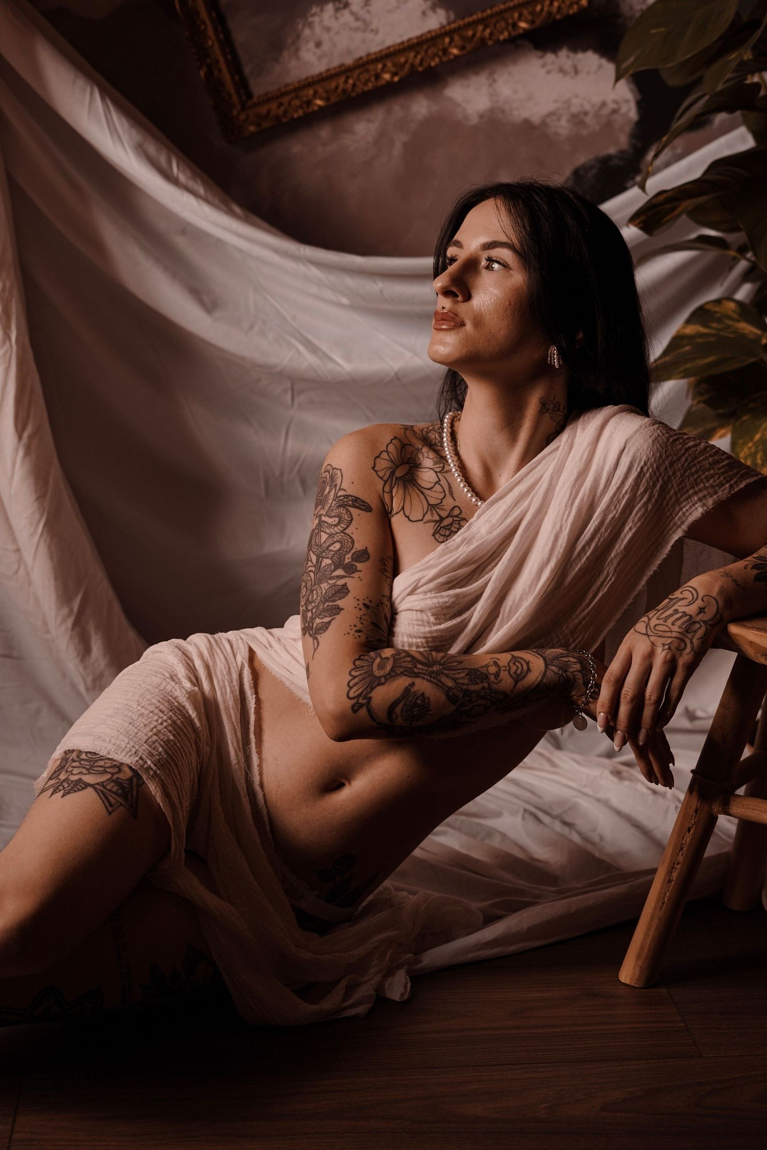 Tattooed brunette woman in sheer wrap dress posing in bohemian studio setting.