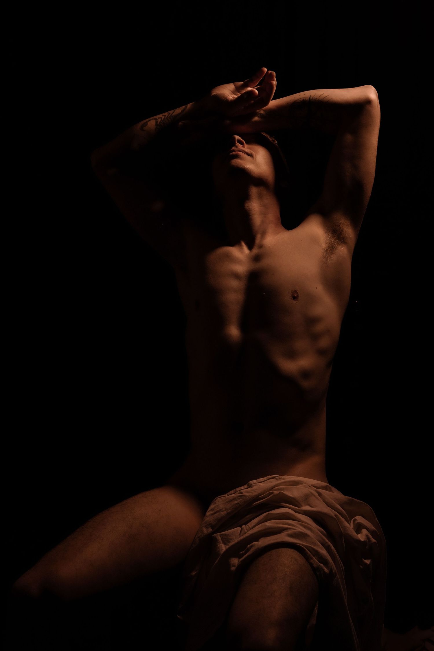 Shirtless muscular man sitting with arms raised overhead in dramatic shadow lighting.