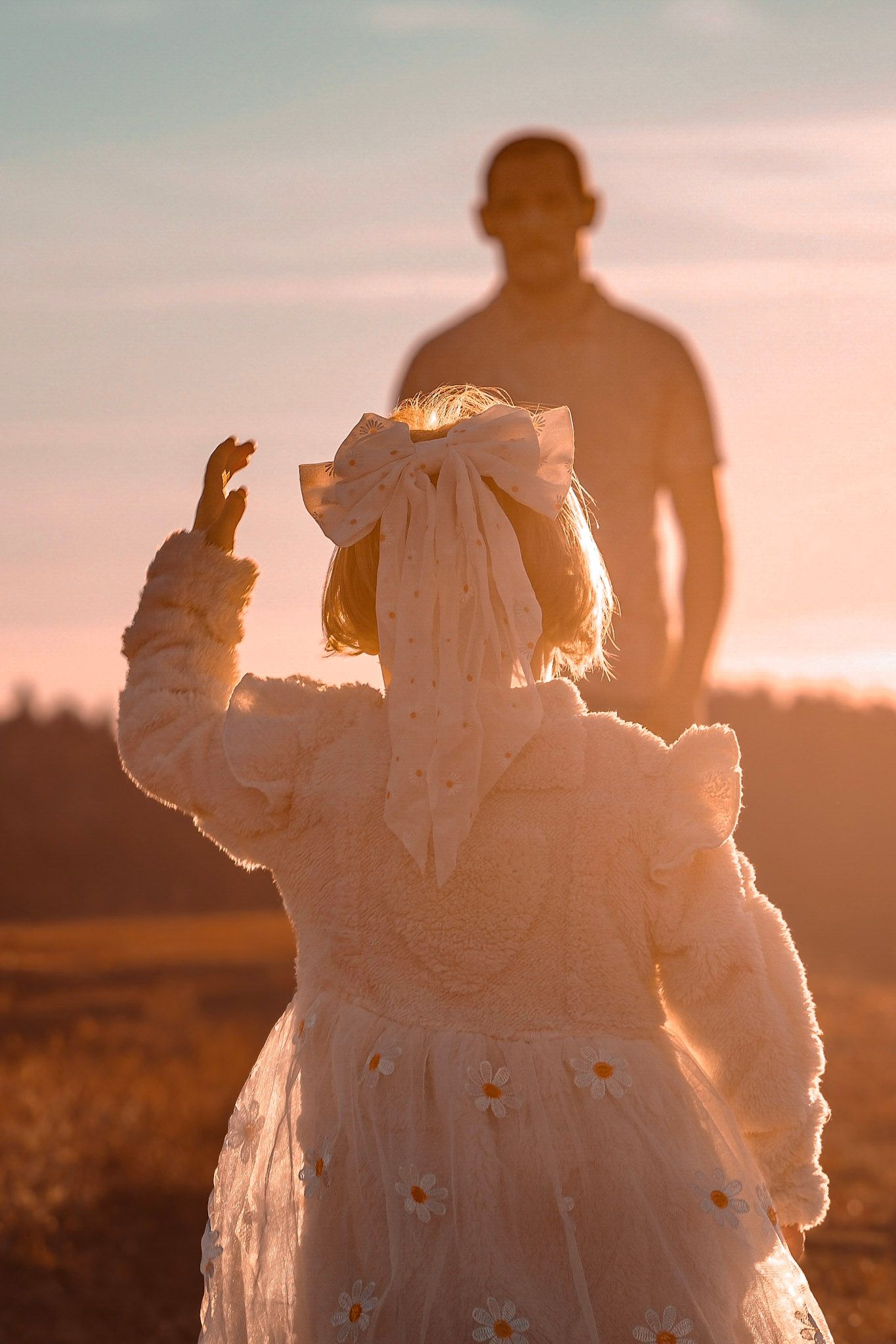 A little girl in a white floral dress reaches toward a man silhouetted against a warm golden sunset in an open field.
