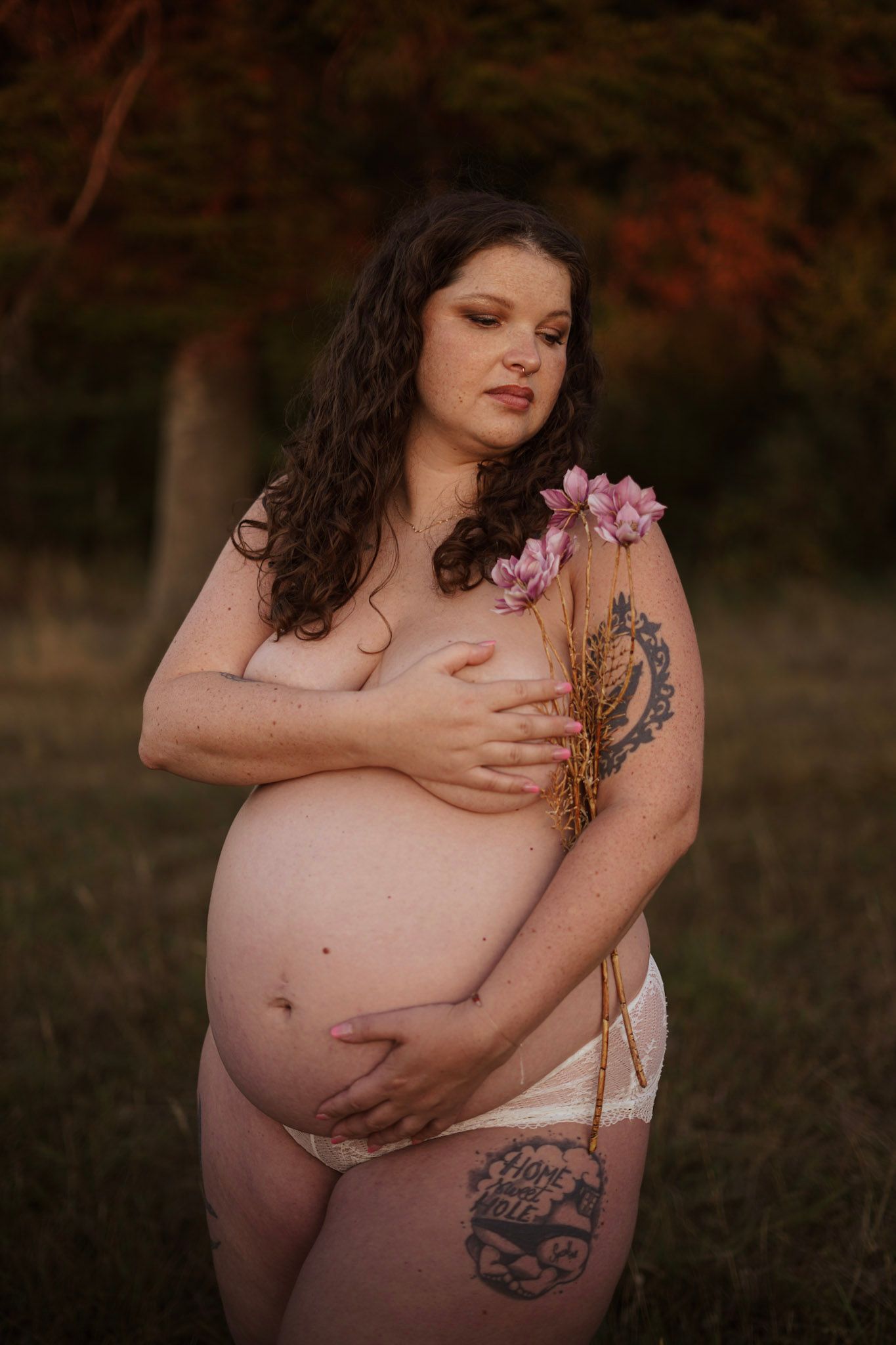 Pregnant woman holding pink flowers outdoors in autumn, showcasing baby bump.