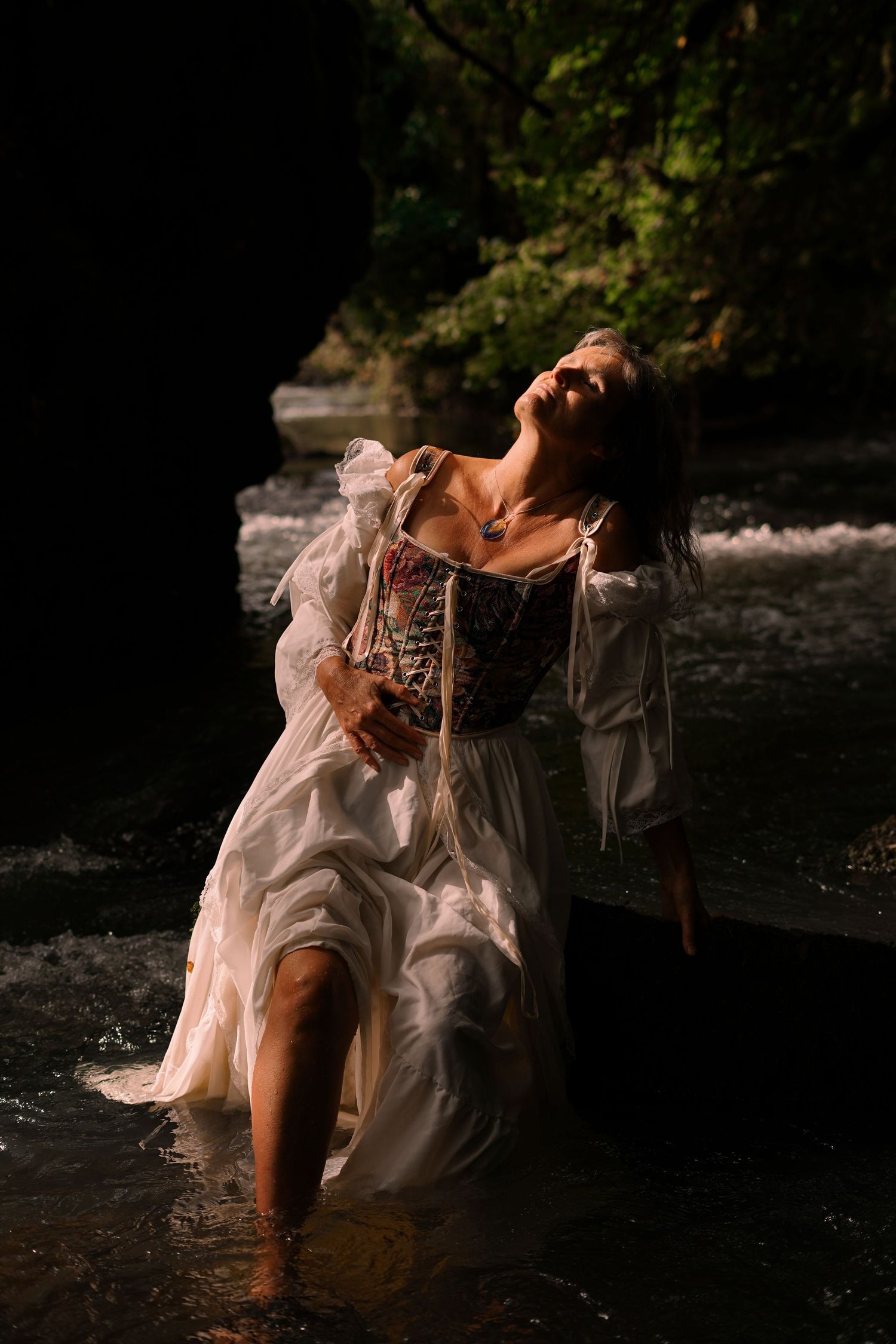 Woman in white dress standing in a river surrounded by lush green forest.
