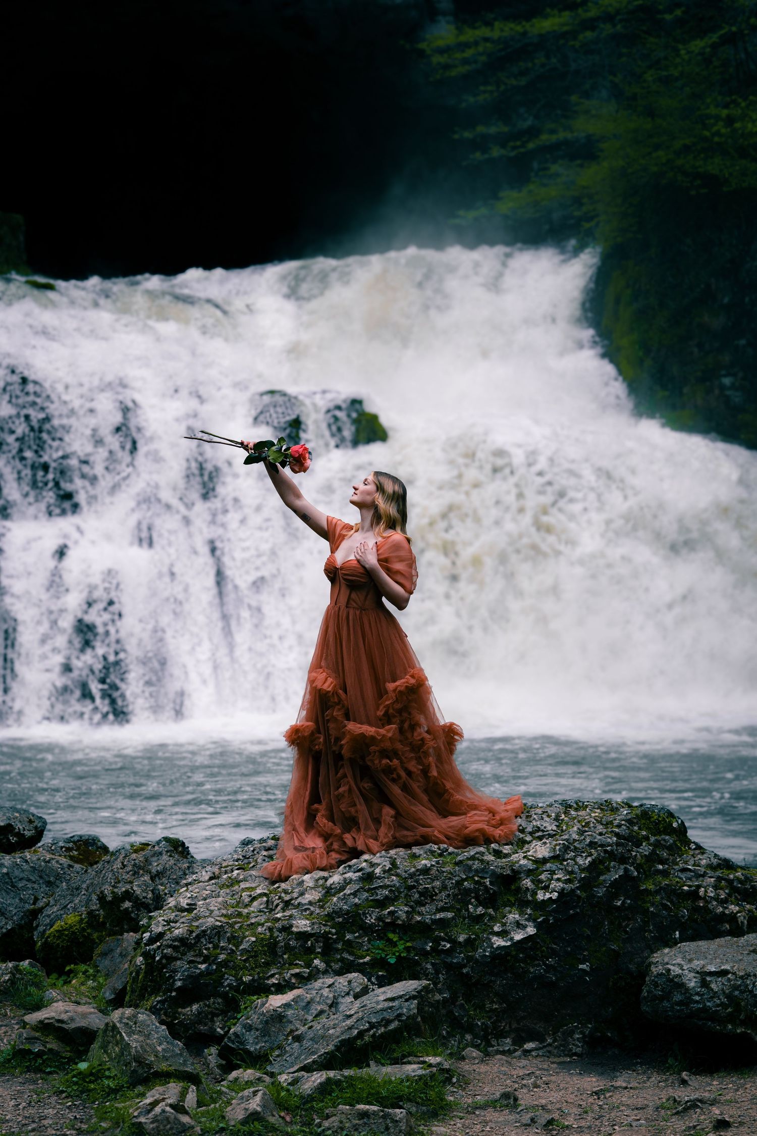 A woman in a flowing rust tulle gown holds a red rose while standing on rocks before a waterfall.