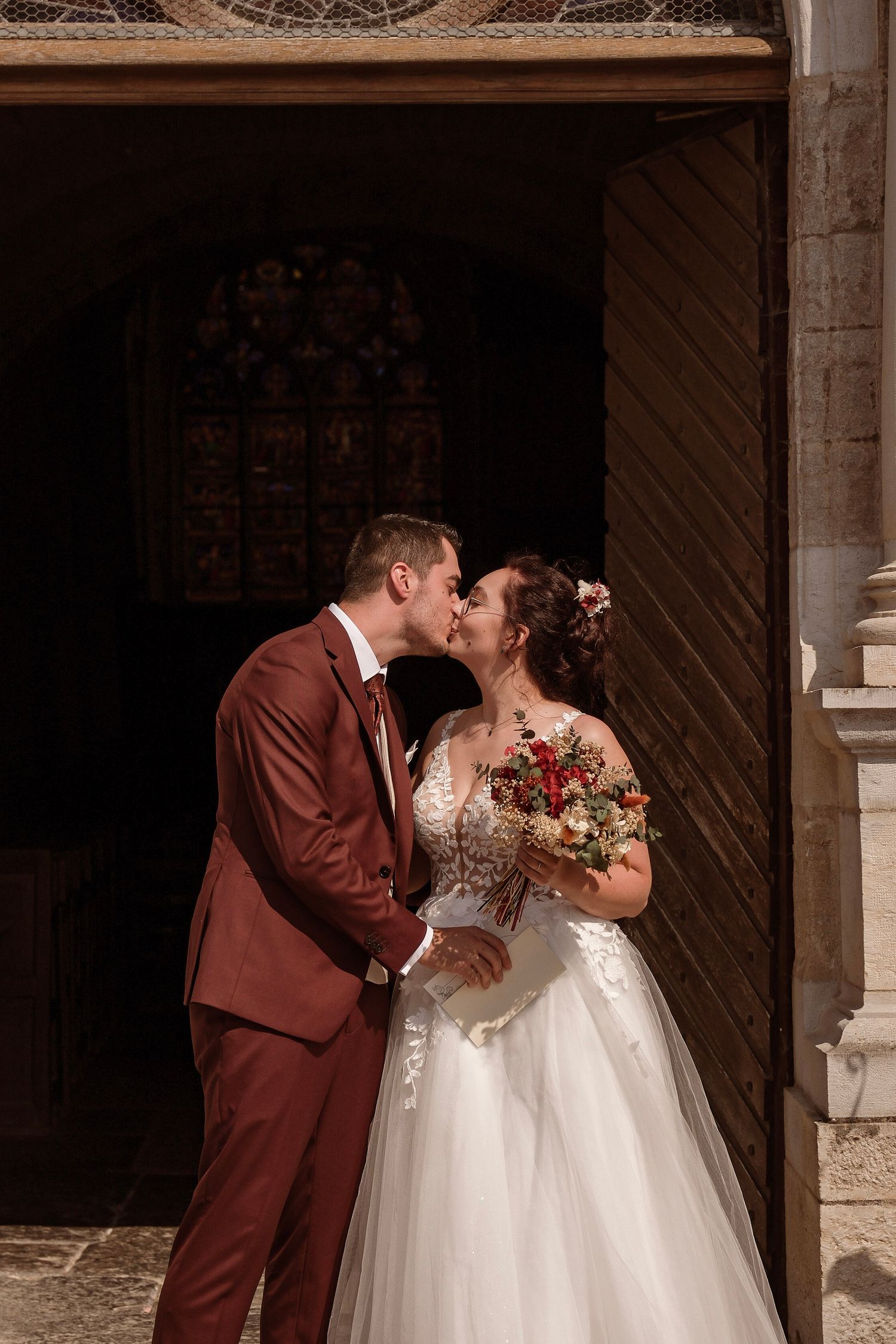 Bride and groom share a kiss at the entrance of a historic church holding a red and white bouquet.