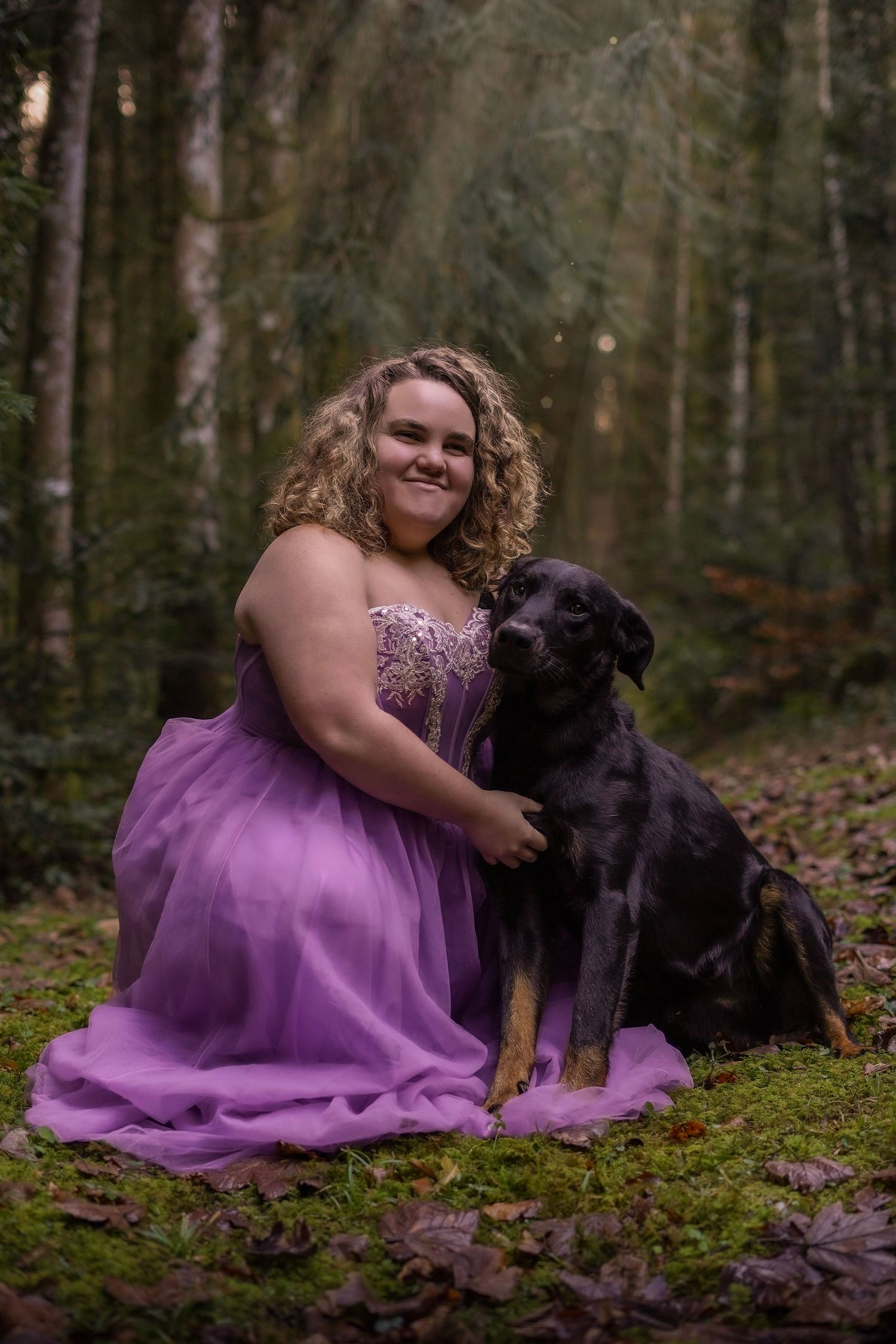 Smiling young woman in purple prom dress posing with black Rottweiler in forest.