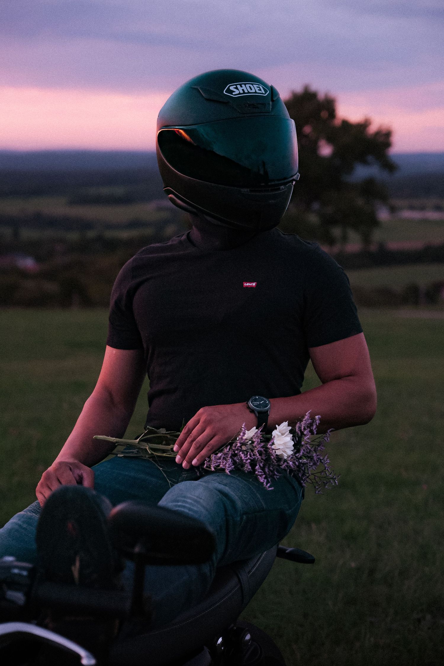 Motorcyclist wearing Shoei helmet holding wildflowers at sunset in an open field.