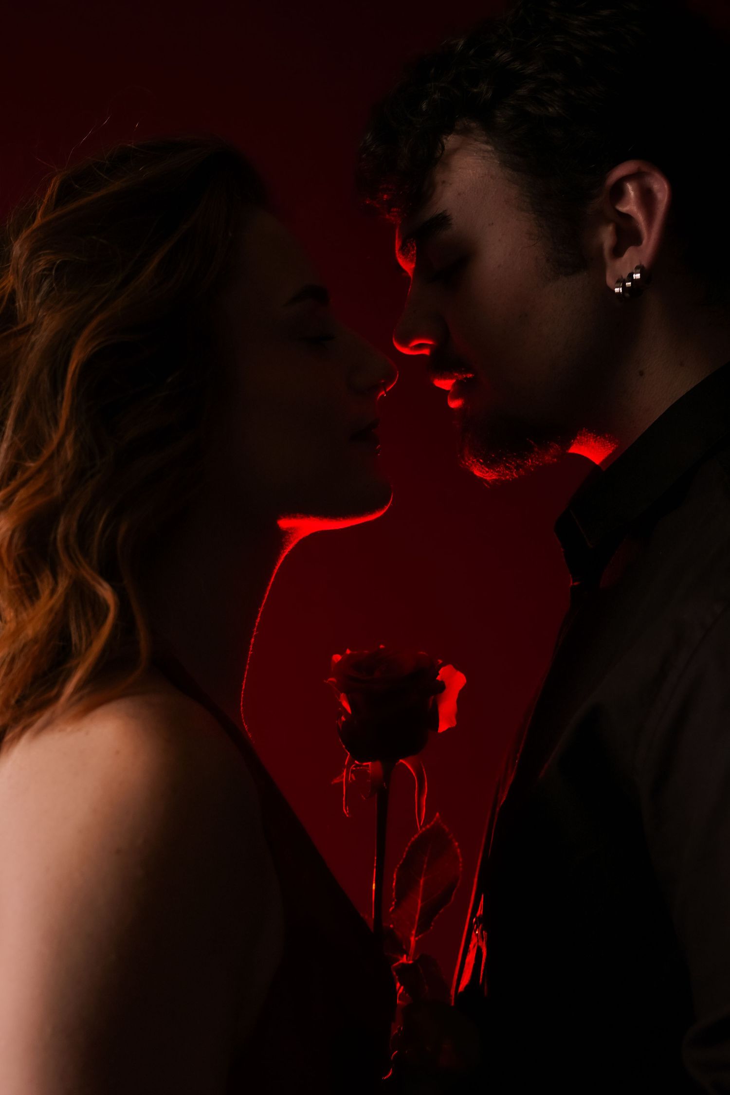 Romantic couple nearly kissing in dramatic red light while holding a single rose.