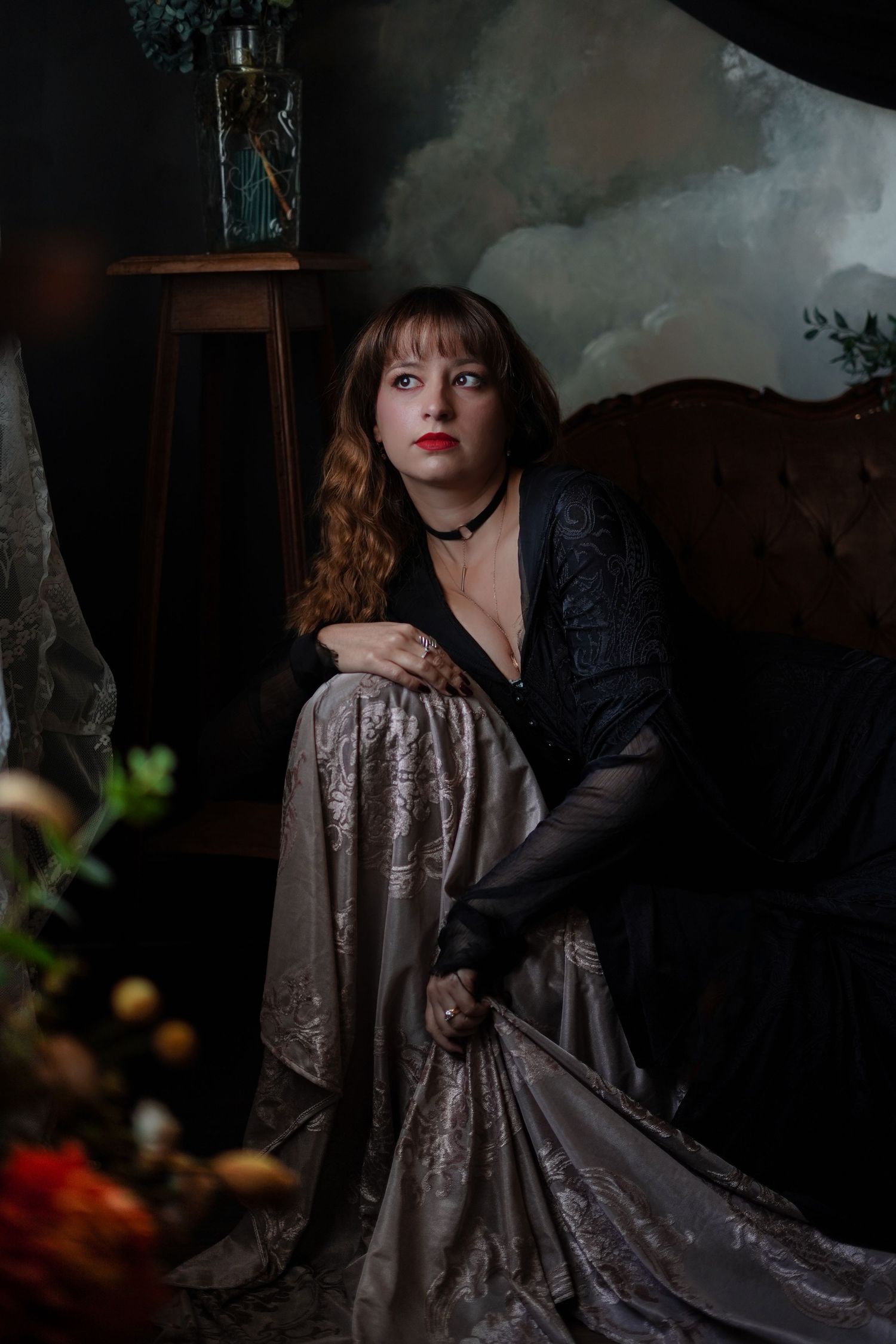 Woman in black lace top and silver skirt seated in dark vintage room.