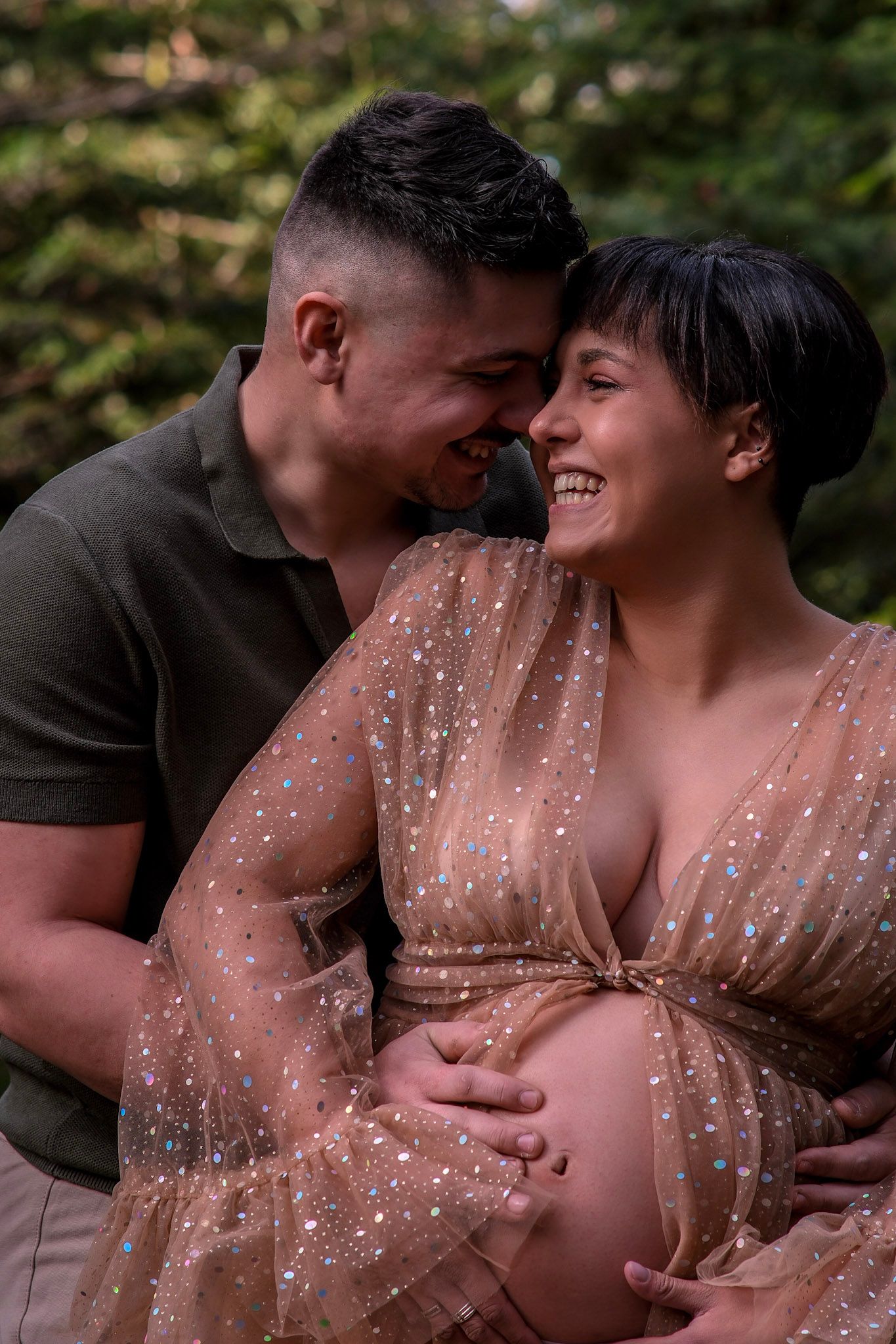 A happy couple shares a tender moment outdoors, with the pregnant woman in a sparkly sheer dress cradling her baby bump.