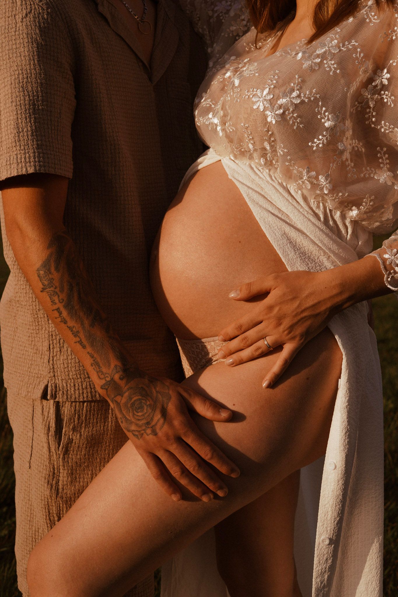 Pregnant woman in white lace robe cradling baby bump outdoors, partner with tattoos embracing her in warm golden light.